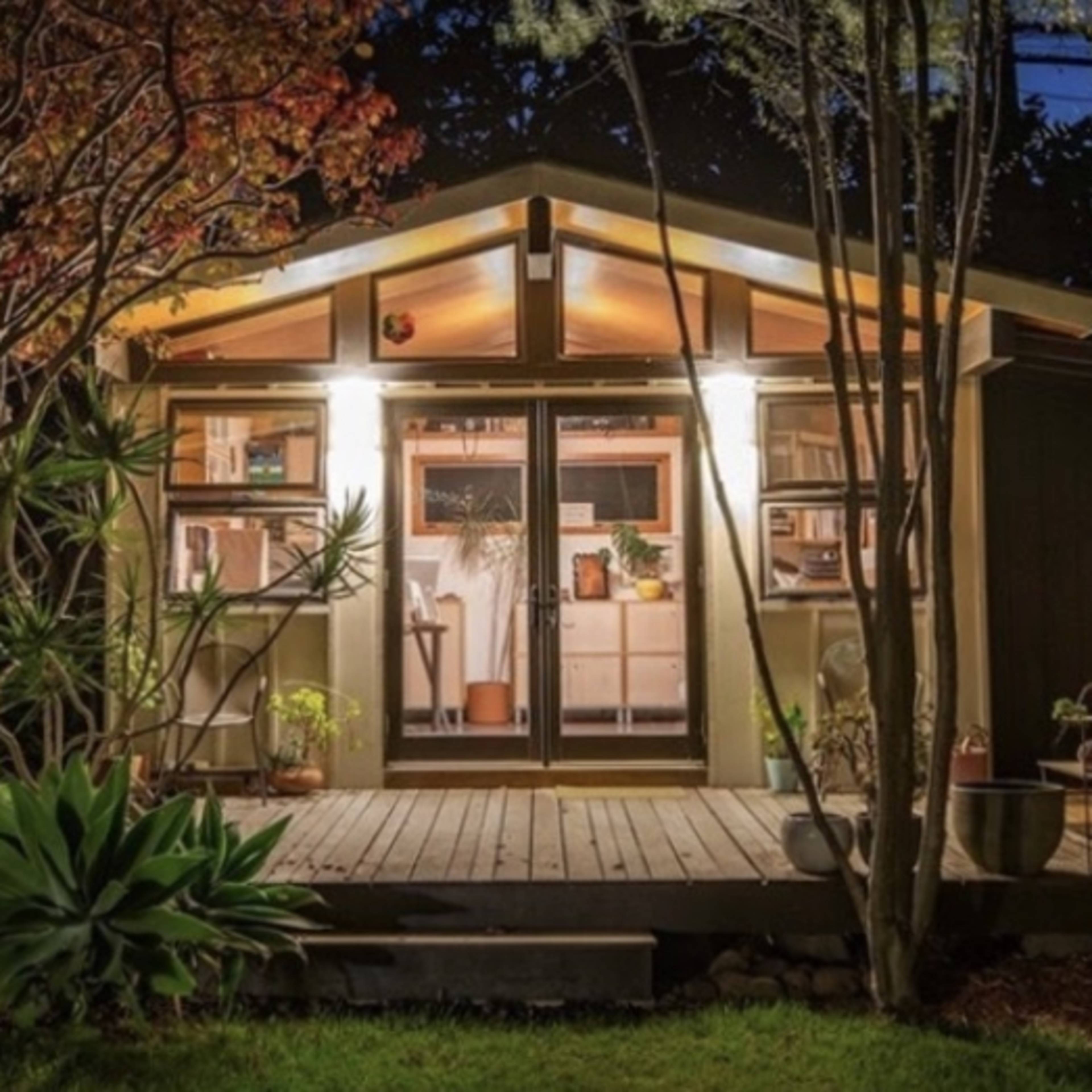 A modern wooden cabin with large windows is illuminated at night, surrounded by lush greenery and a wooden deck.