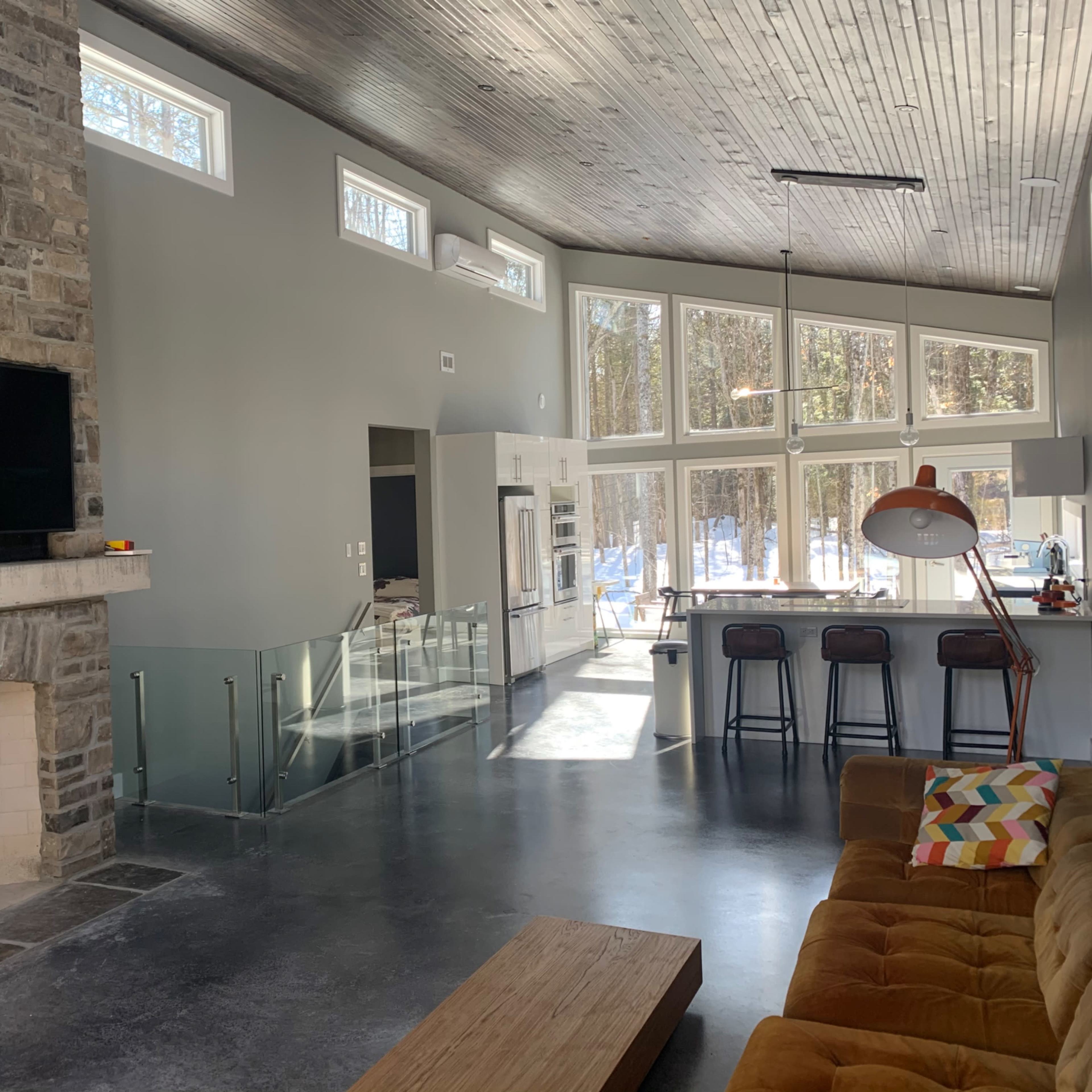 A modern living room featuring a stone fireplace, large windows with views of a wooded area, and a contemporary seating arrangement.