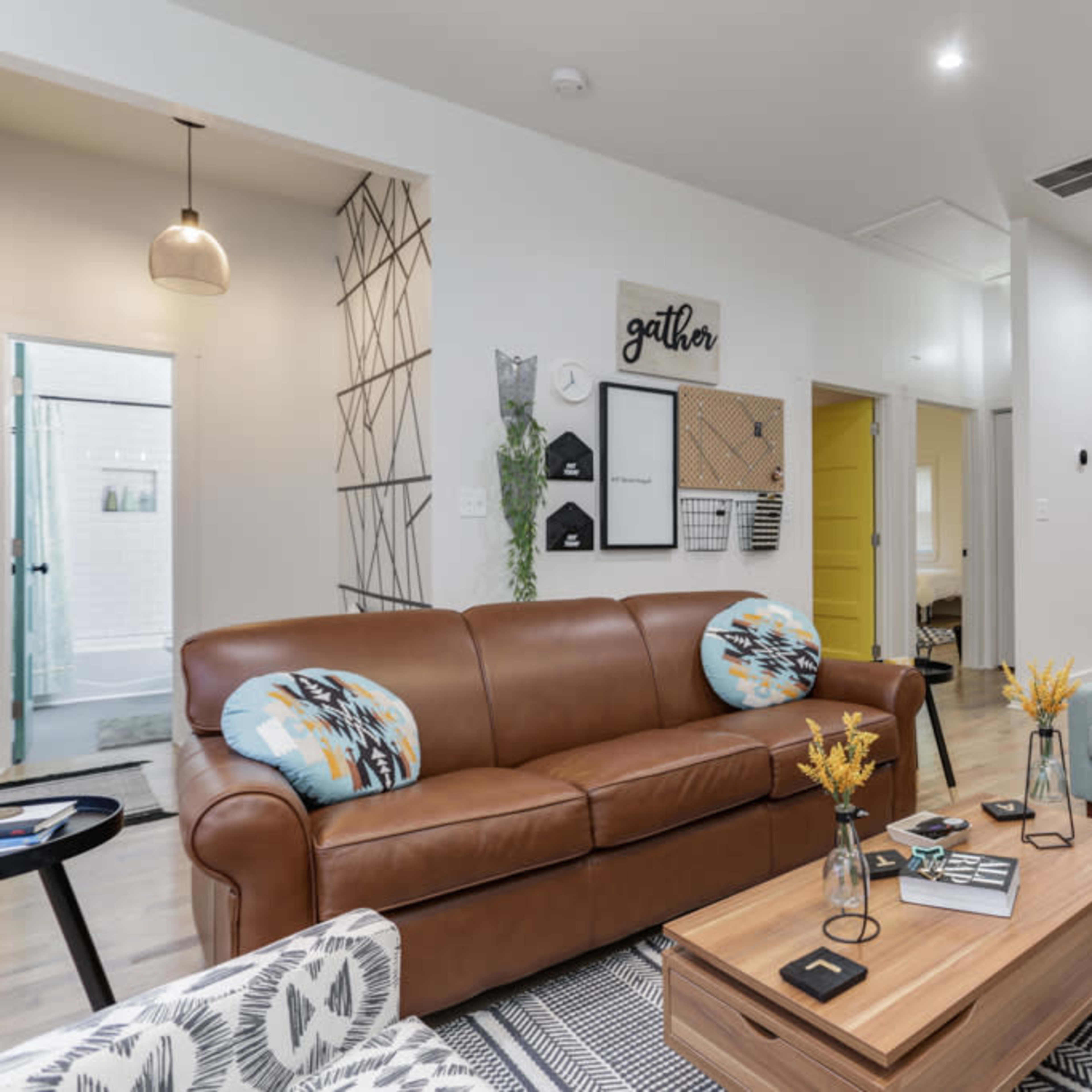 A modern living space with a brown leather sofa, blue accent chairs, a wooden coffee table, and a dining area in the background featuring multiple framed pictures on the wall.