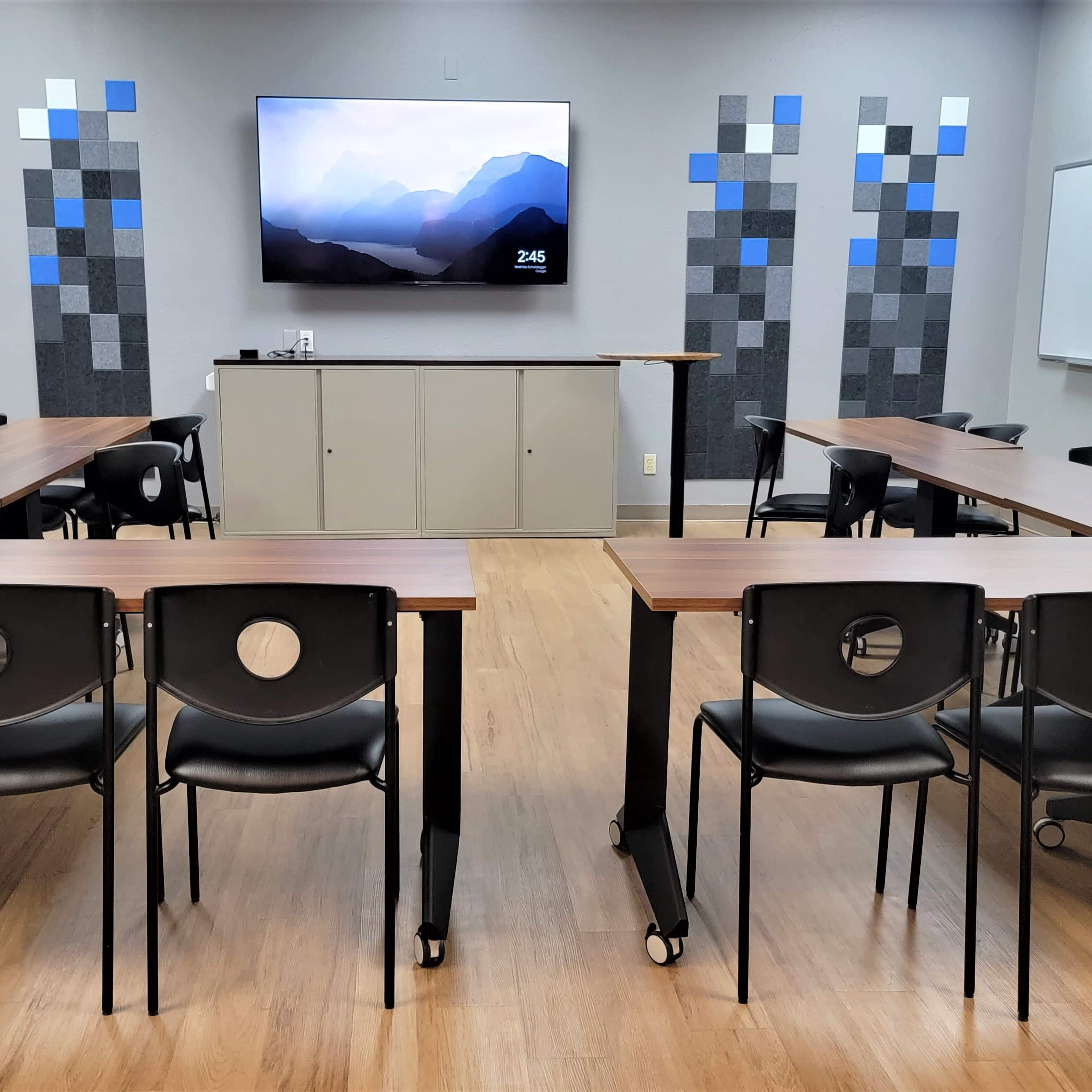 A classroom with movable tables arranged in a U-shape, featuring black chairs, a wall-mounted television, and patterned gray and blue walls.