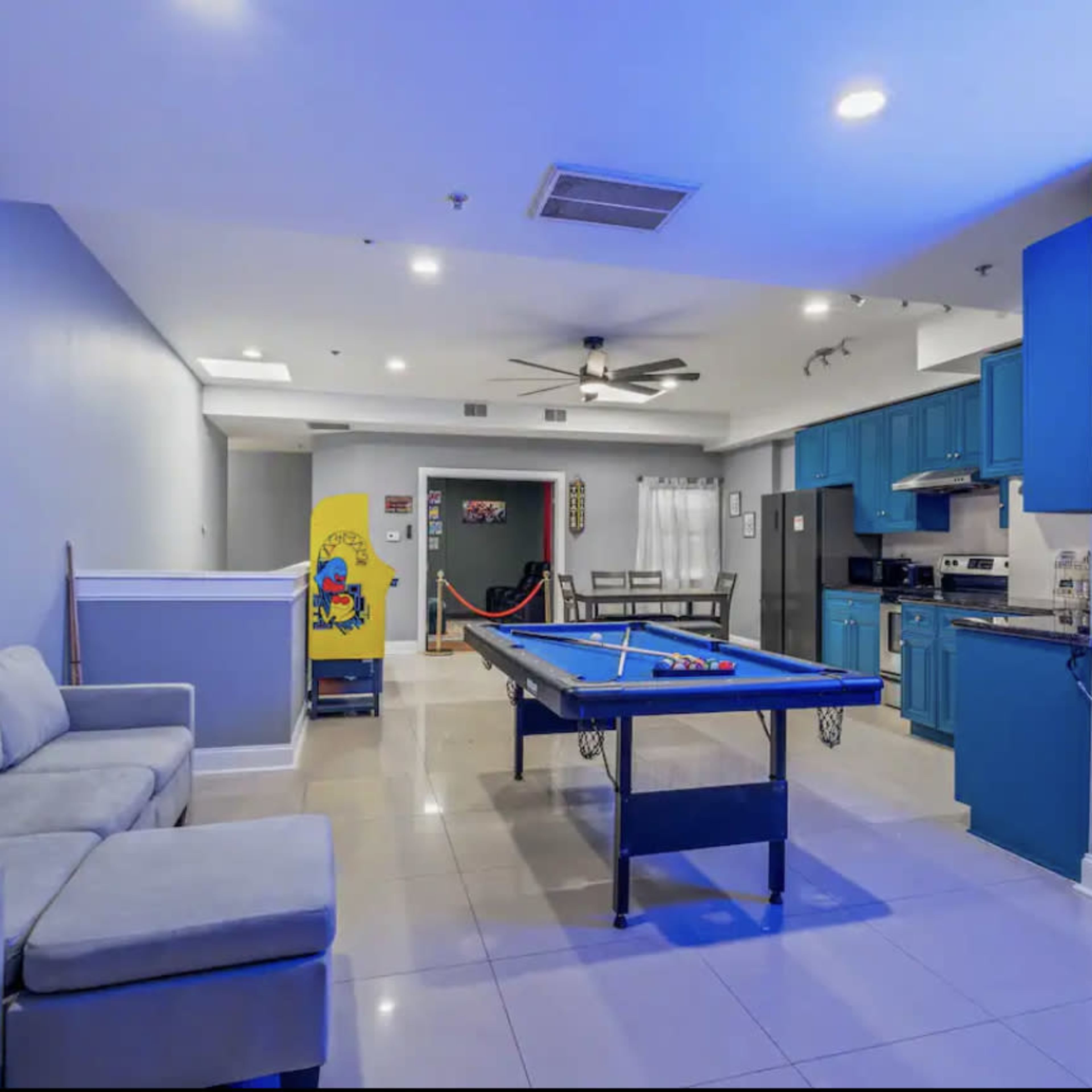A modern game room with a pool table, a couch, and a neon sign, featuring a kitchen area in the background.