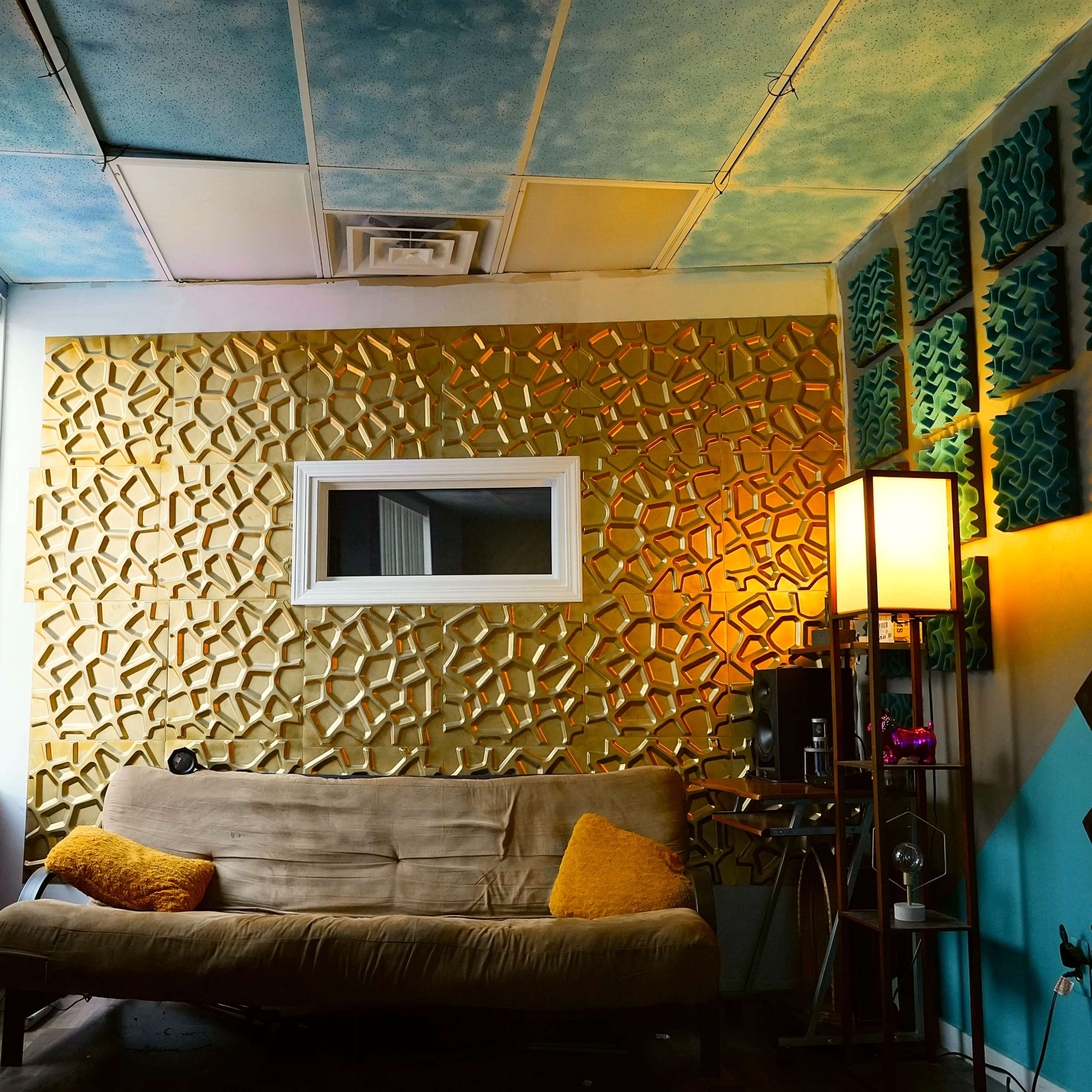 A small room featuring a textured gold accent wall, a beige couch with an orange cushion, and colorful acoustic paneling on the side walls.