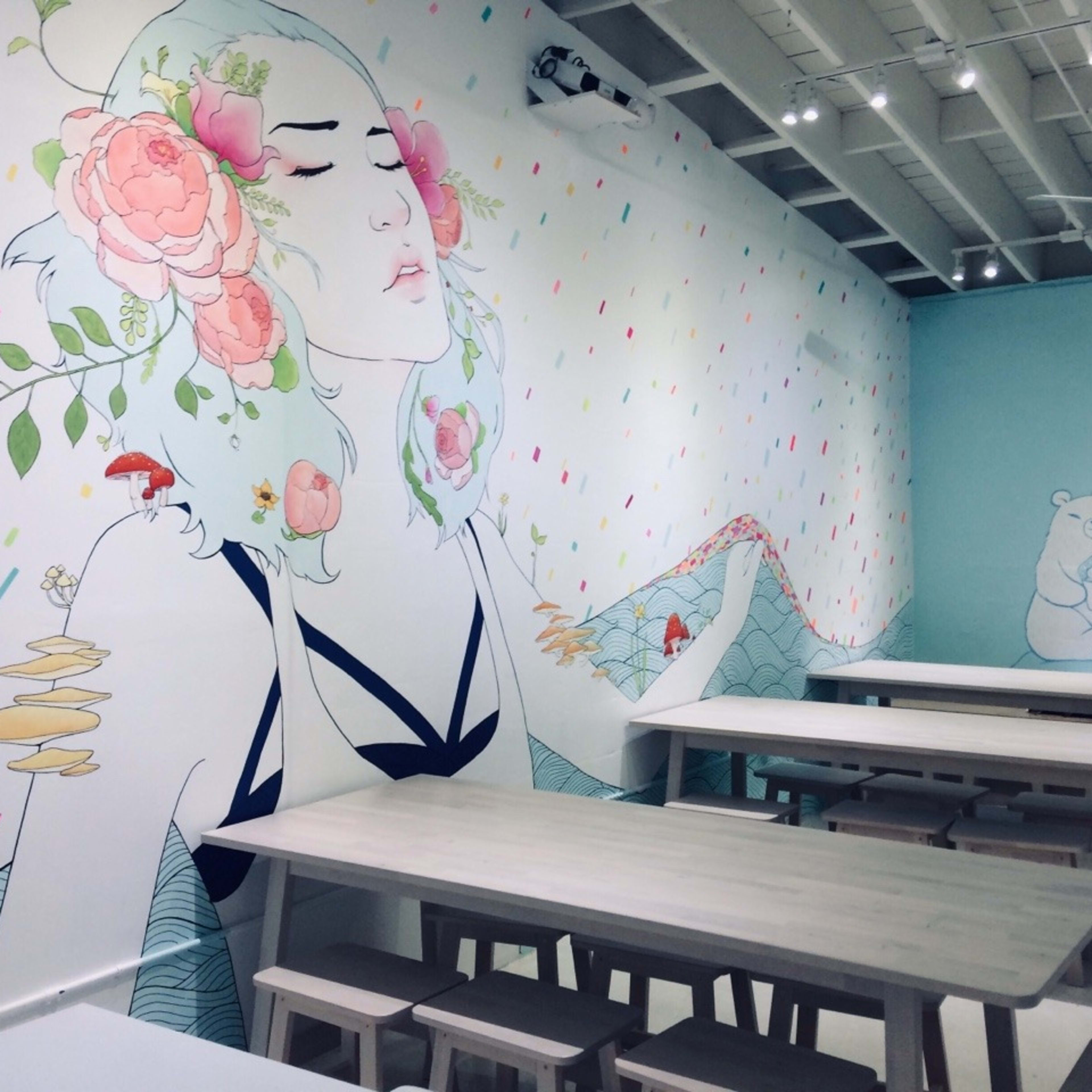 A dining area with wooden tables and chairs, featuring a mural of a woman with floral elements and a bear on the wall.