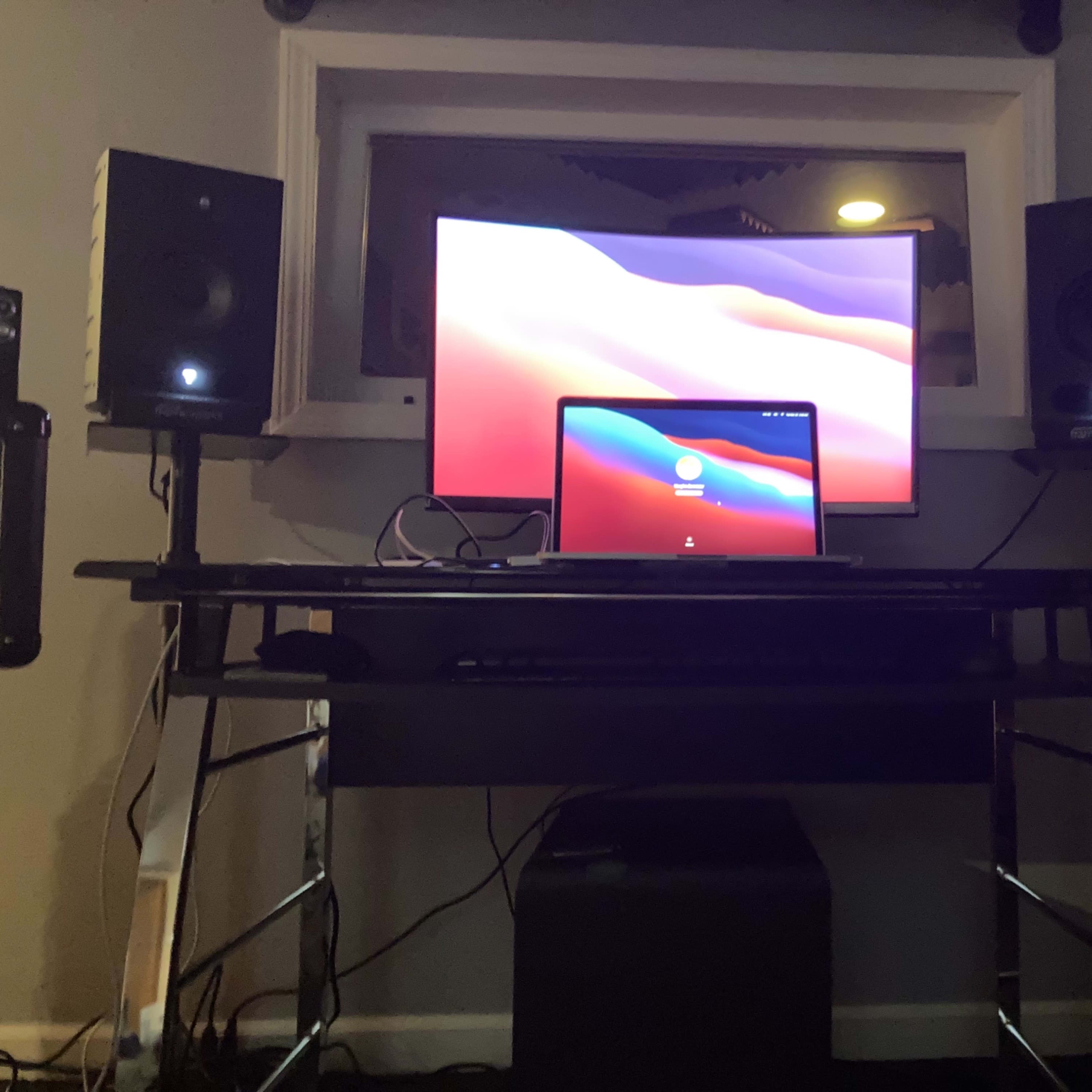 A desk setup with a laptop and a larger monitor, flanked by two speakers, in a room with dim lighting.