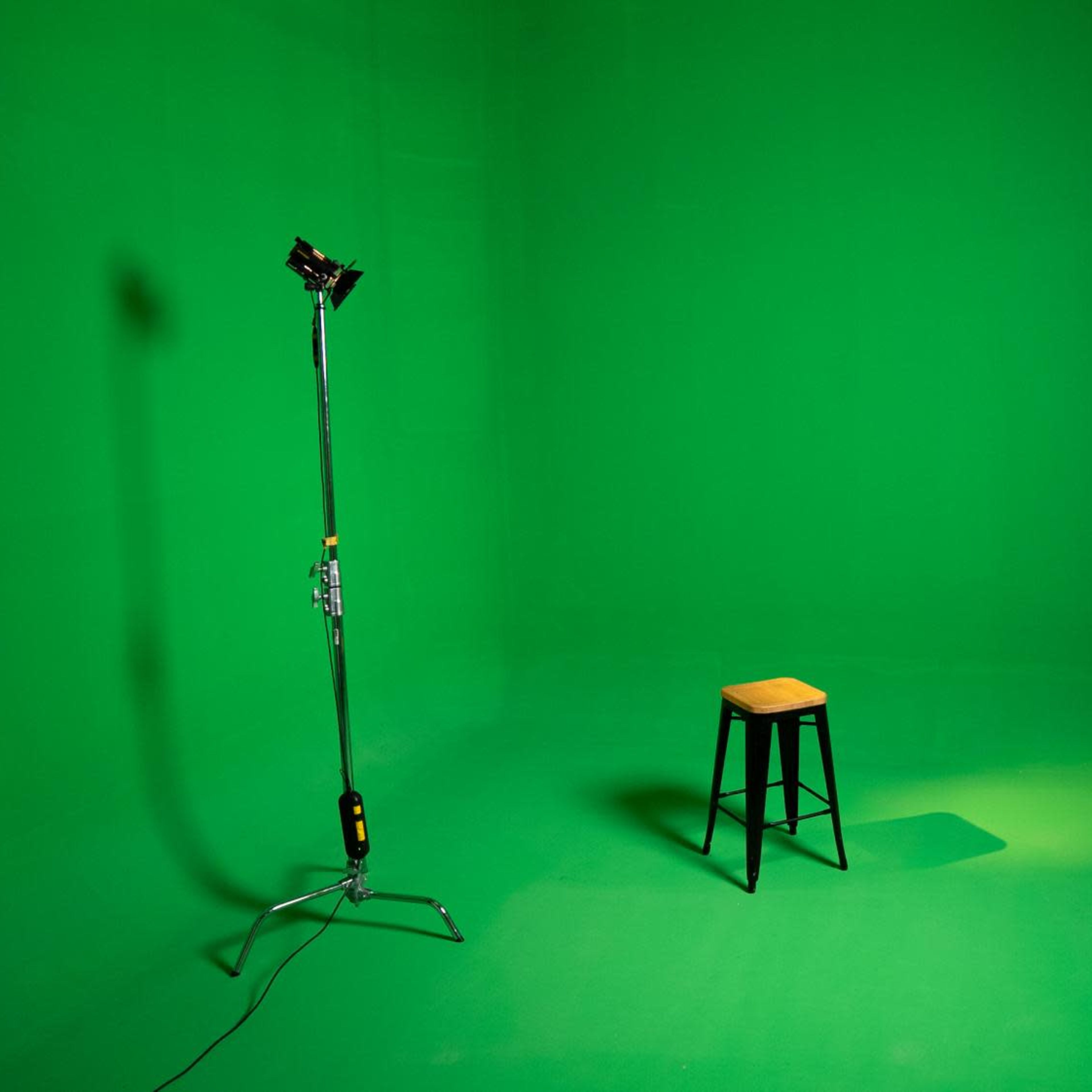 A green screen studio setup featuring a single wooden stool, a light stand, and a camera on a tripod.