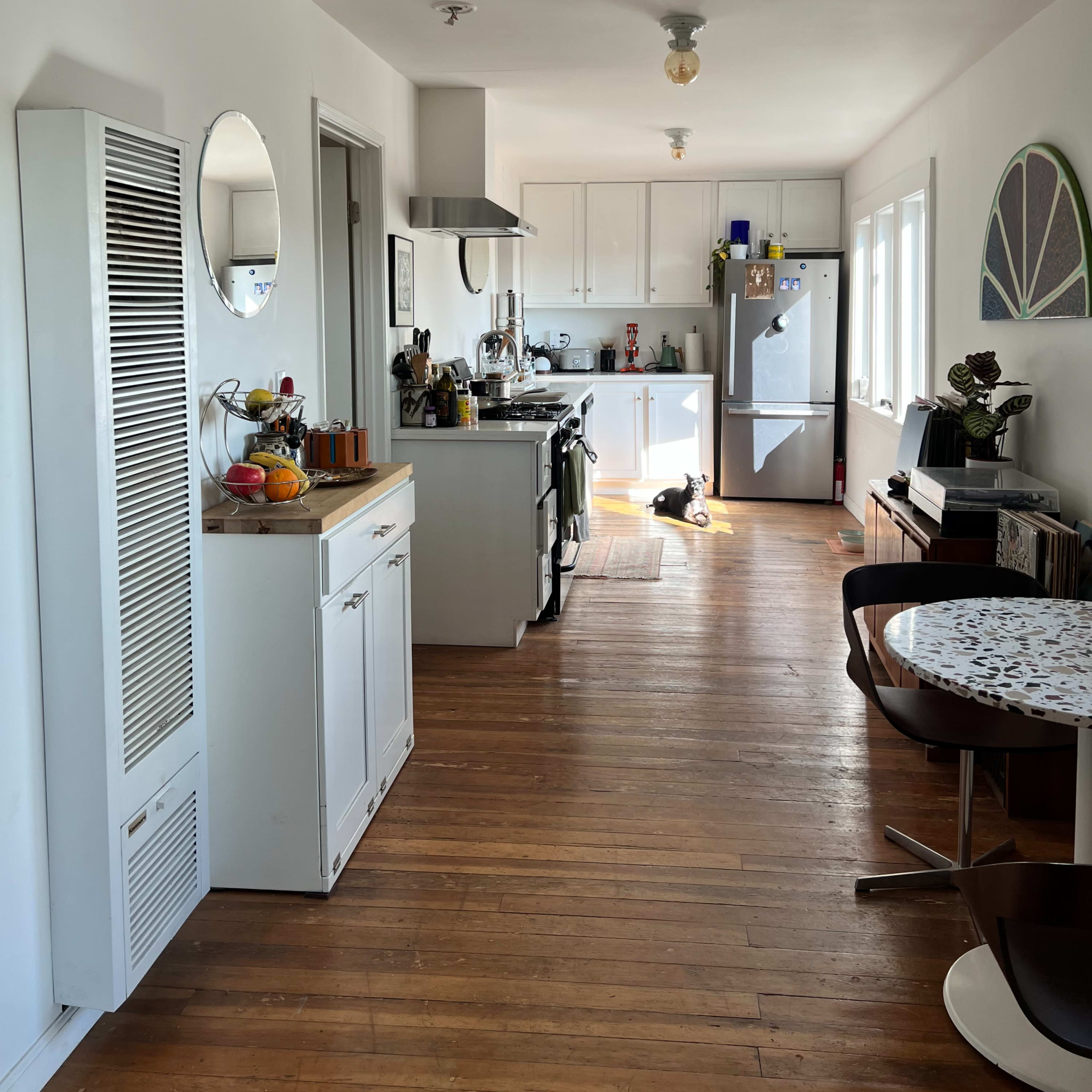 A bright kitchen with wooden floors features a stove, refrigerator, and dining area with a round table.