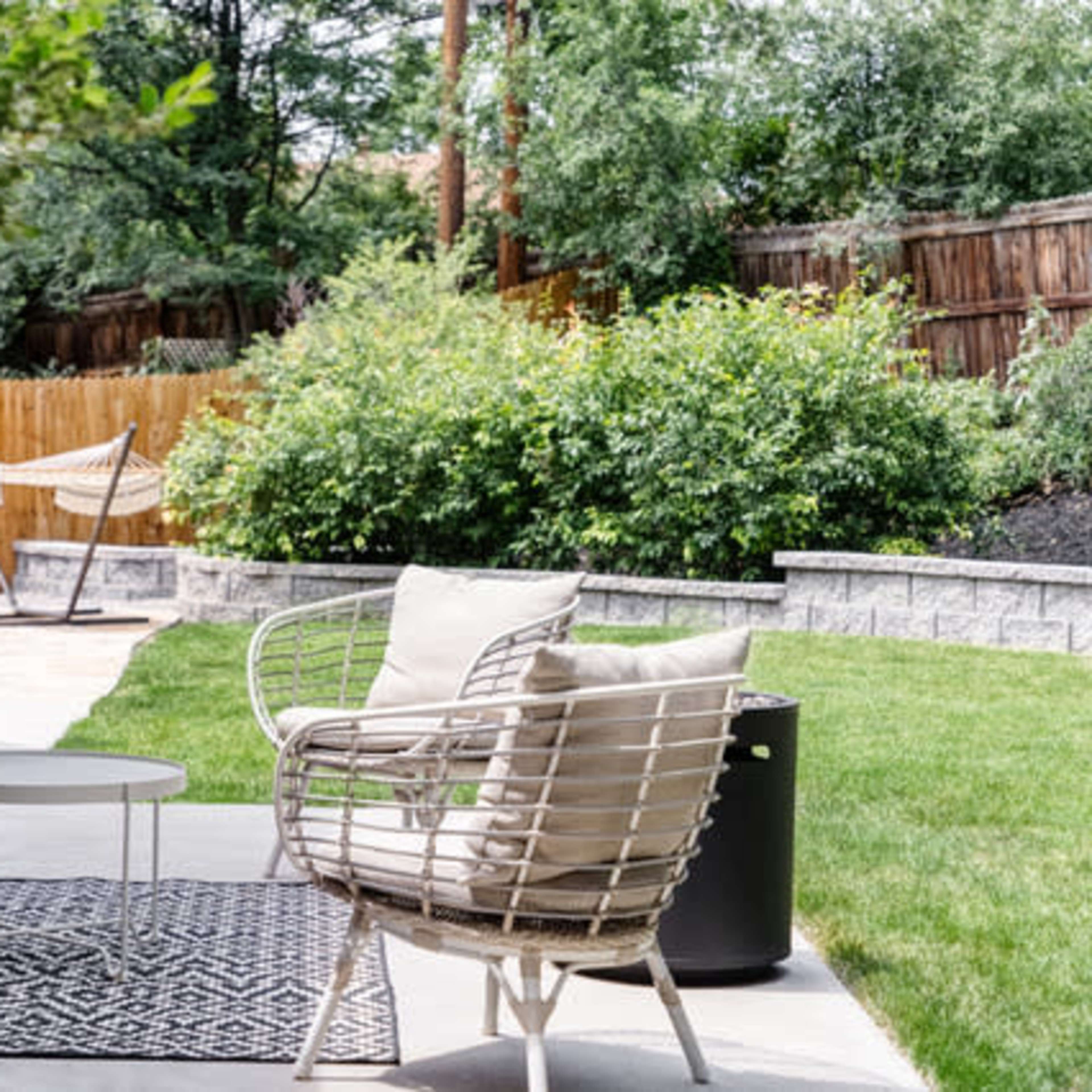A patio area features a white chair with cushions, a small round table, and a neatly manicured lawn surrounded by greenery and a wooden fence.