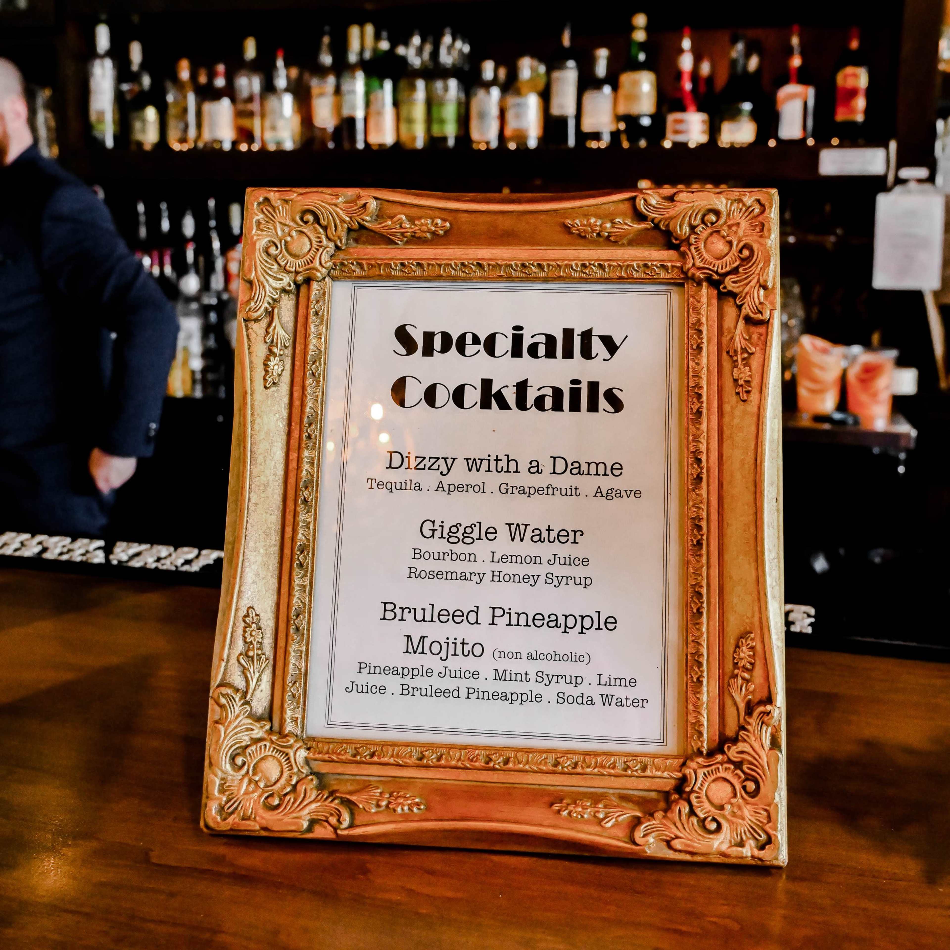 A decorative framed menu displaying specialty cocktails is placed on a bar counter in front of a shelf filled with various liquor bottles.