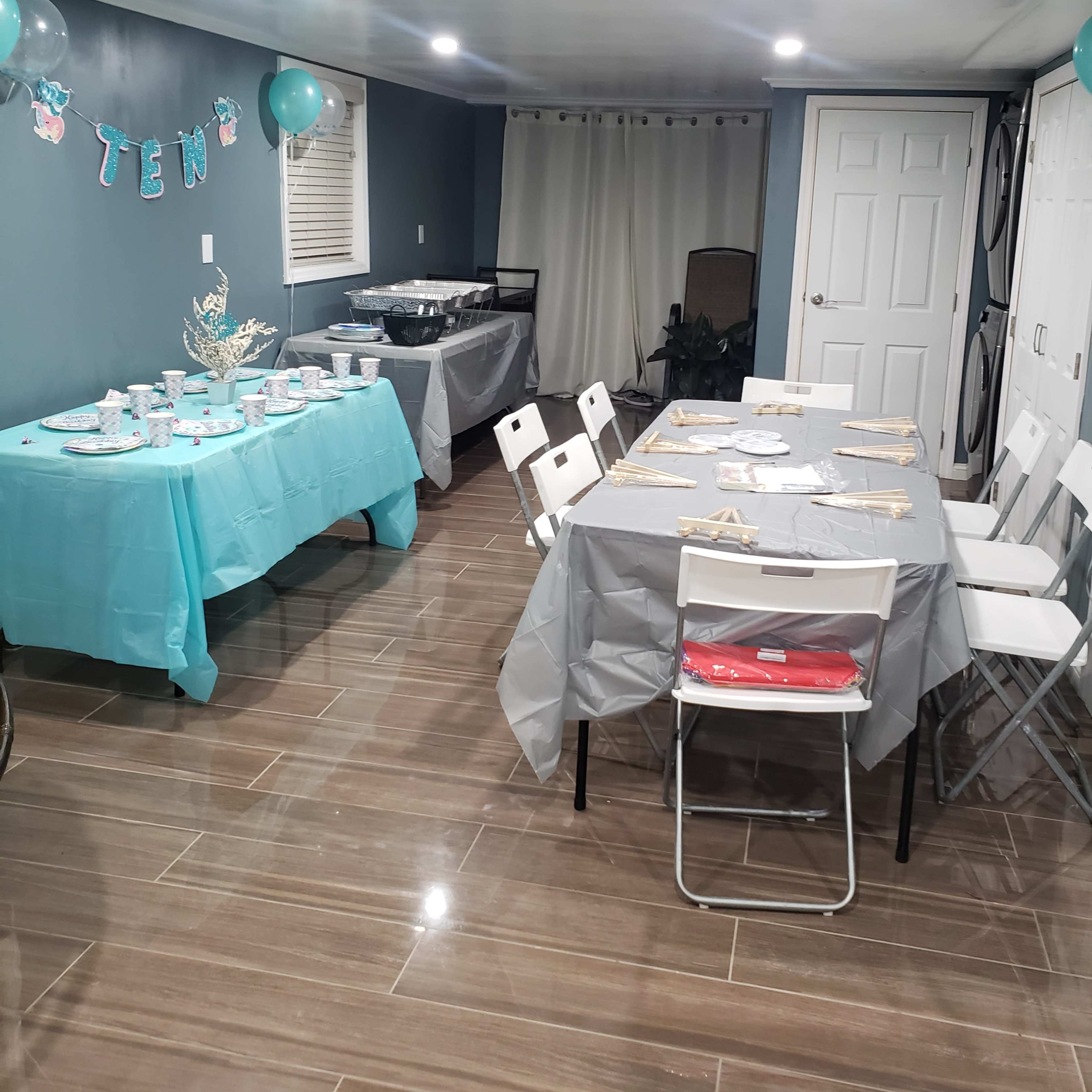 A party setup in a basement with tables covered in light blue and gray tablecloths, decorated with balloons and banners.