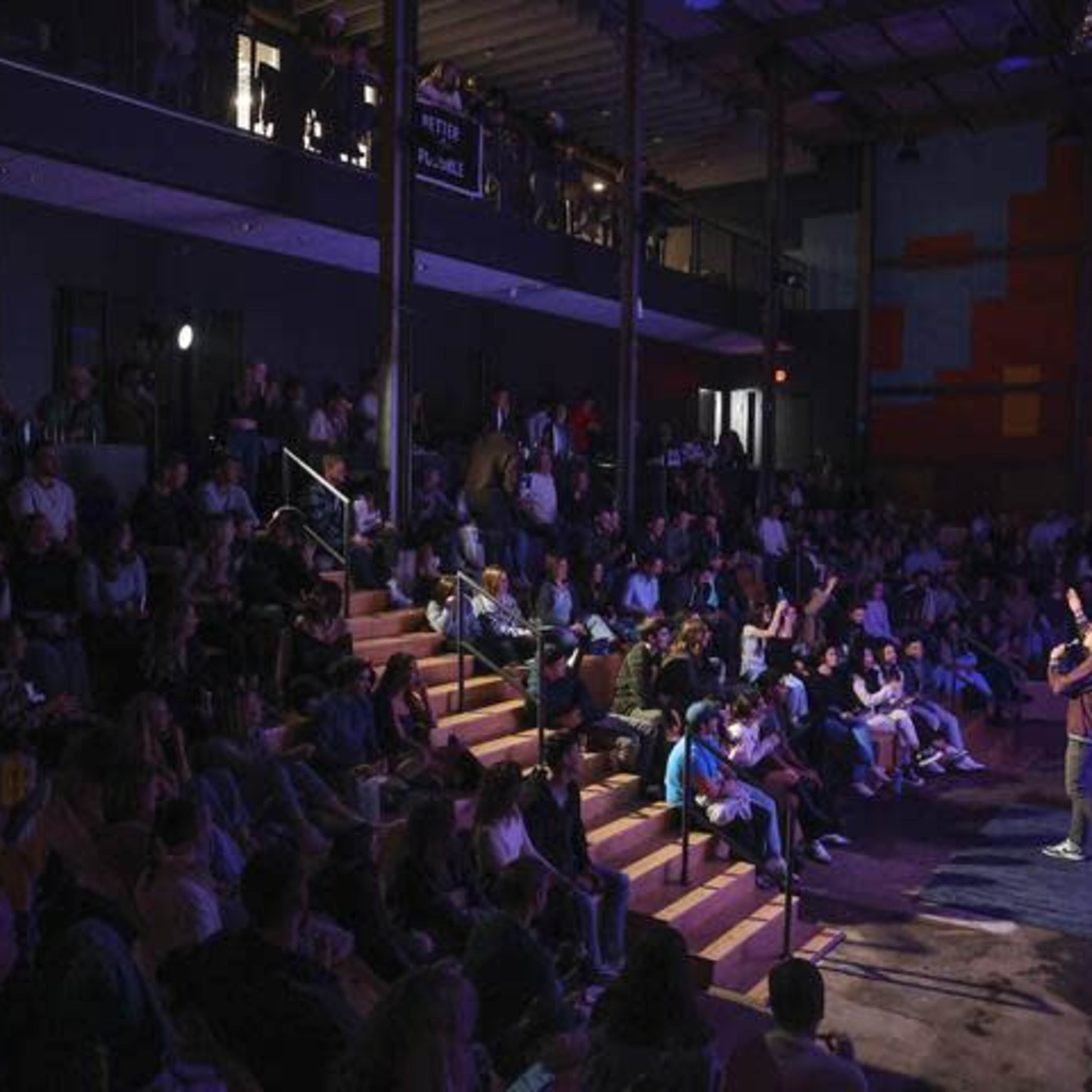 A speaker stands on a stage addressing a large audience seated in a theater-style venue with multiple levels.
