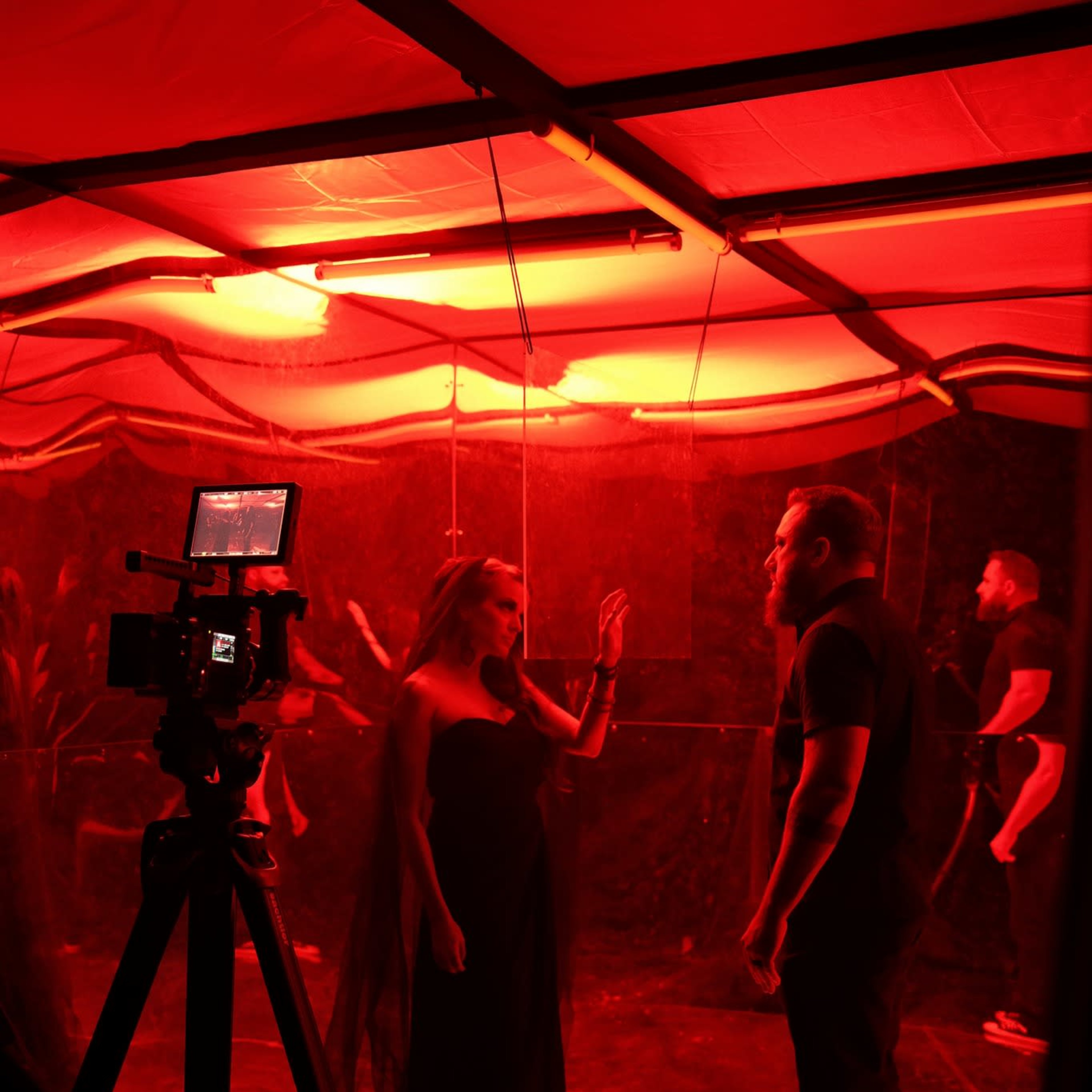 A woman in a black dress gestures toward a man in a black shirt under red lighting in a partially draped room.
