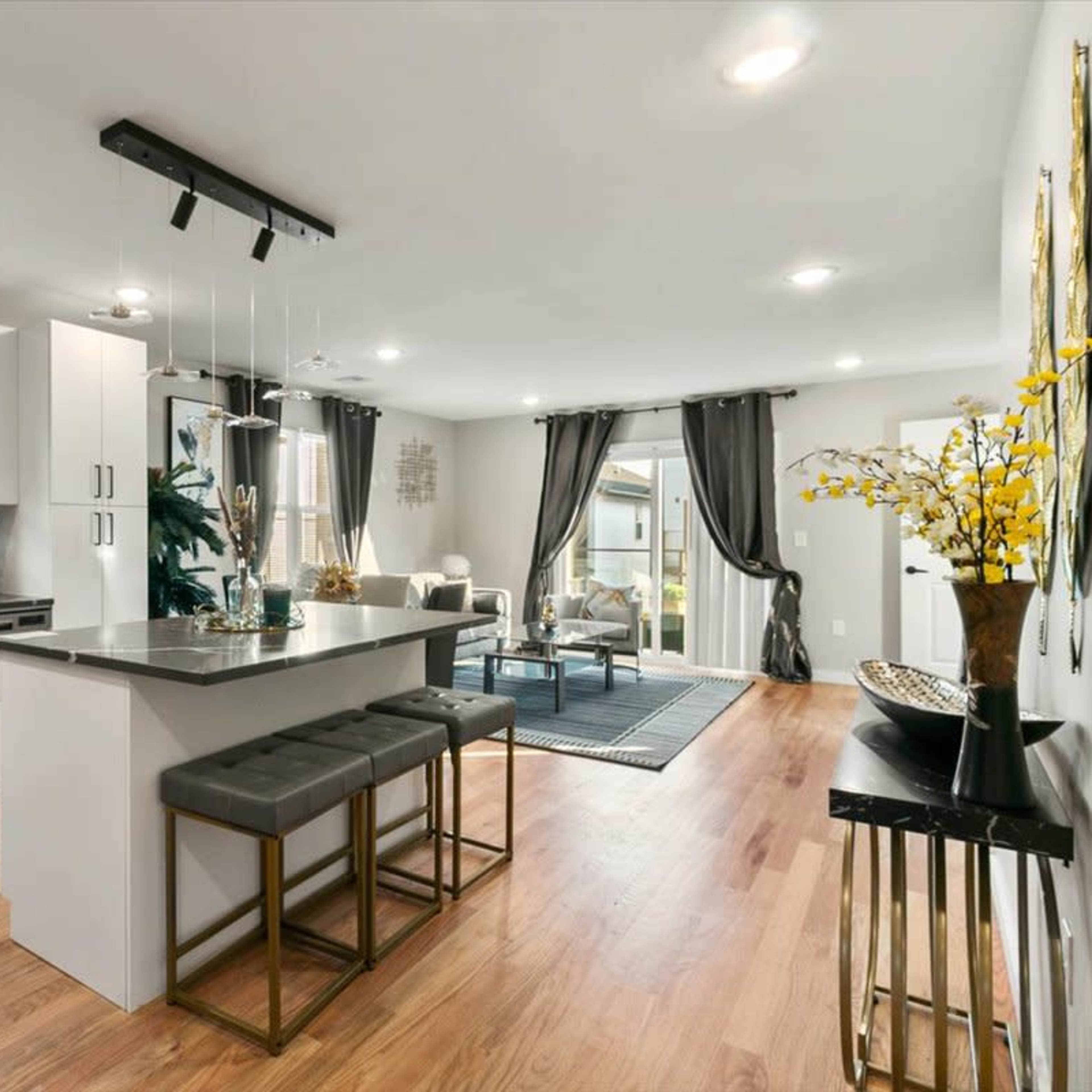 A modern kitchen and living area feature a central island with seating, sleek white cabinetry, and large windows that open to a patio.