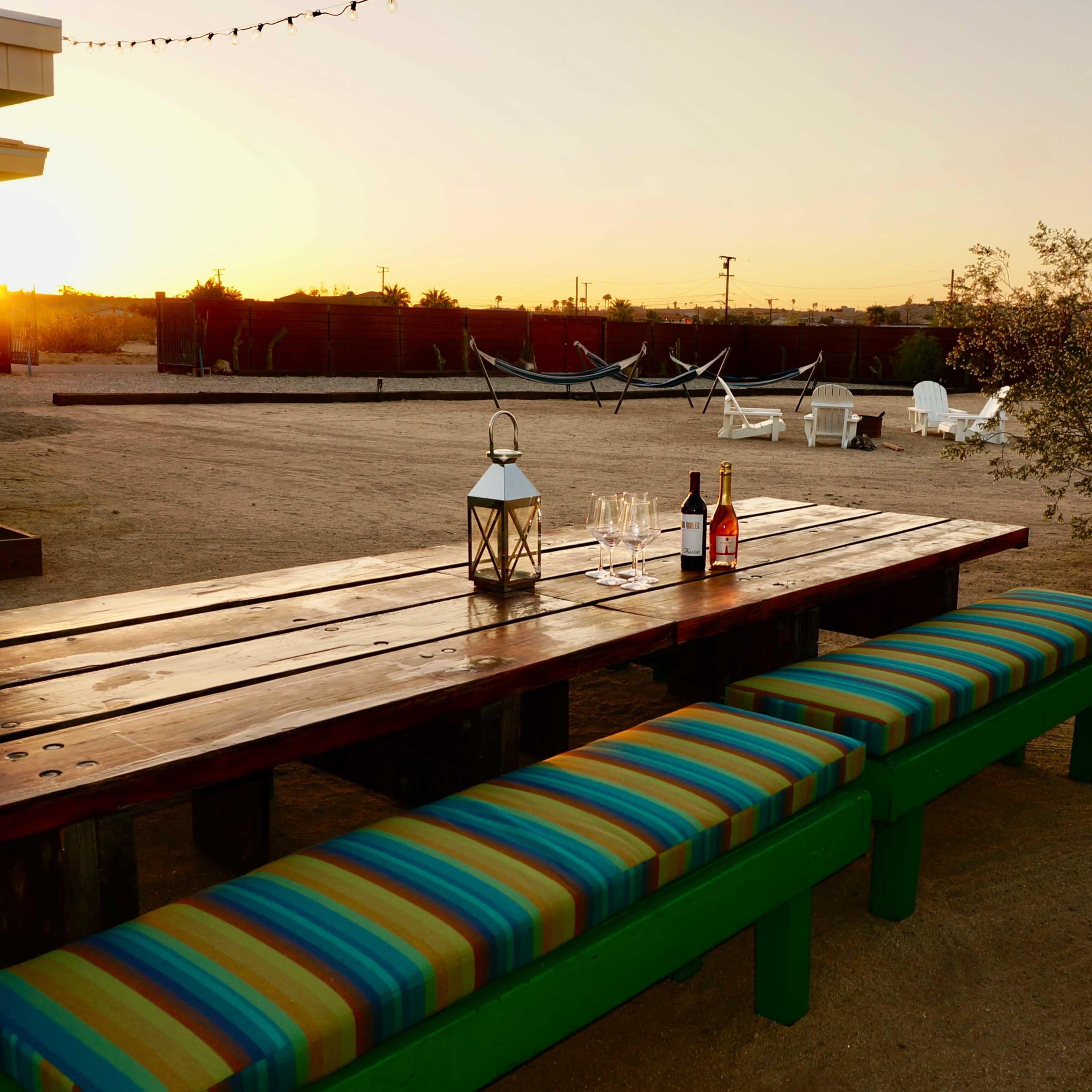 A long wooden table with colorful benches sits in a dusty outdoor area as the sun sets in the background.