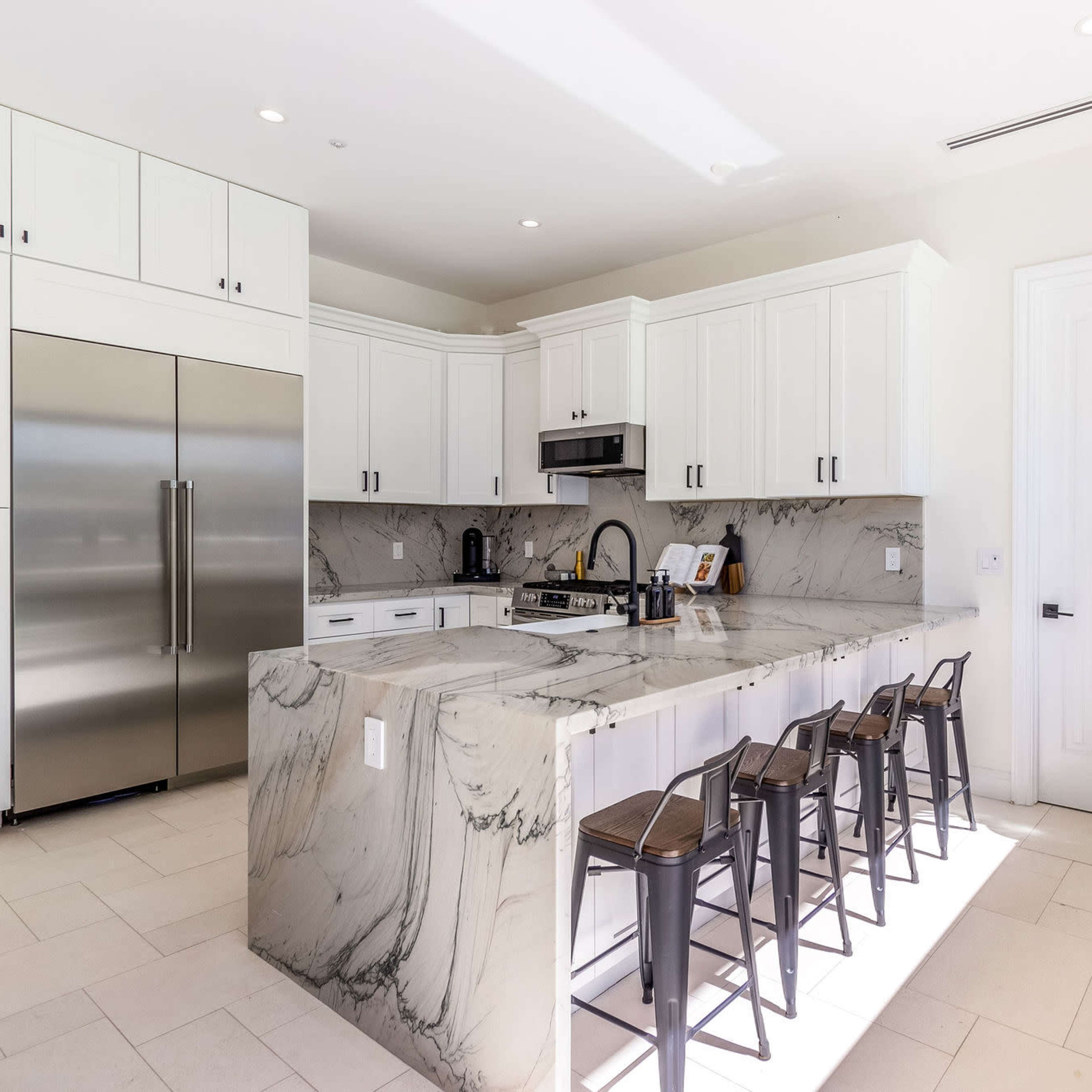 A modern kitchen features white cabinetry, a large marble countertop, stainless steel appliances, and seating for four at the island.