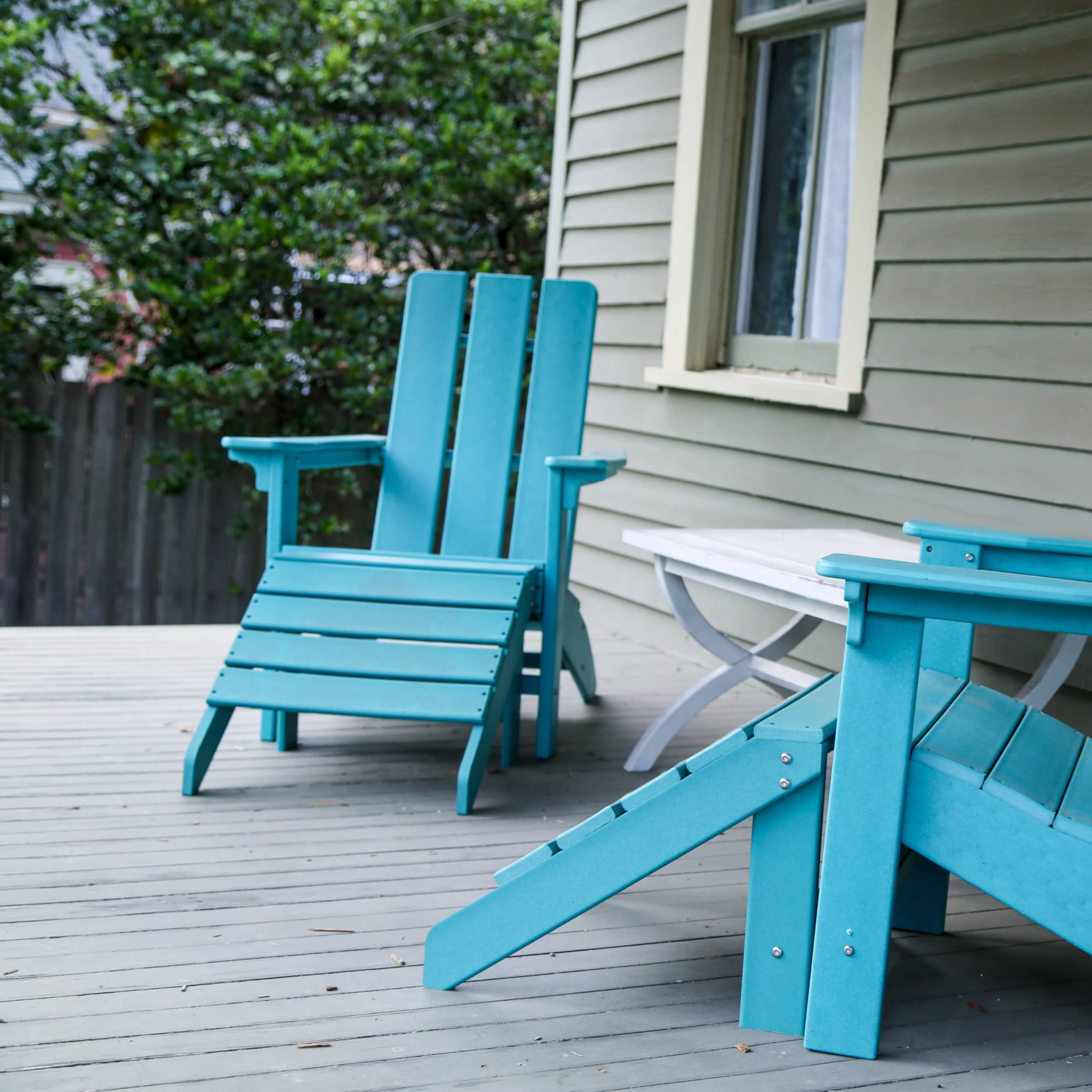 Two turquoise Adirondack chairs sit beside a white side table on a wooden porch with green siding.