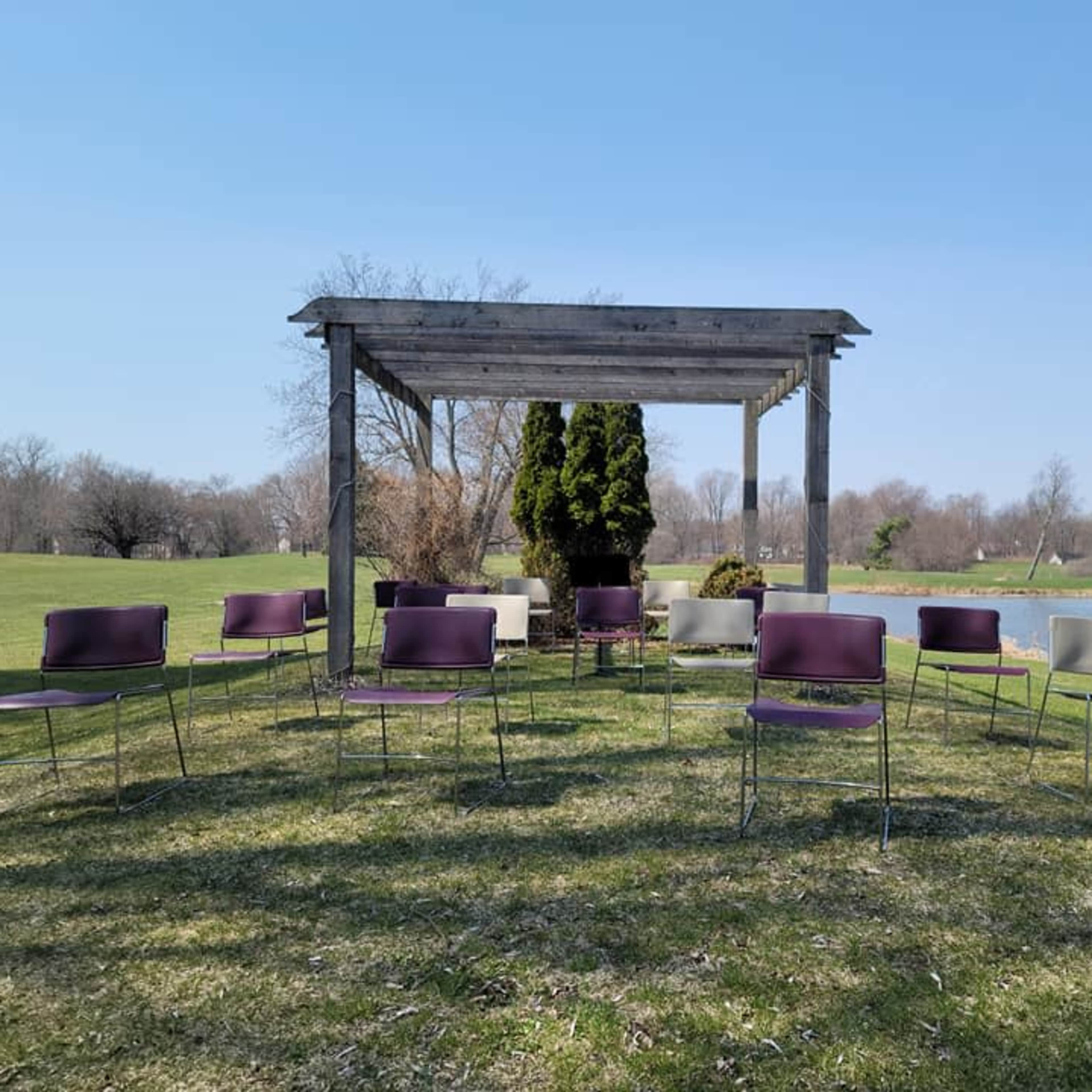 A circle of chairs surrounds a wooden pergola in a grassy area near a body of water.