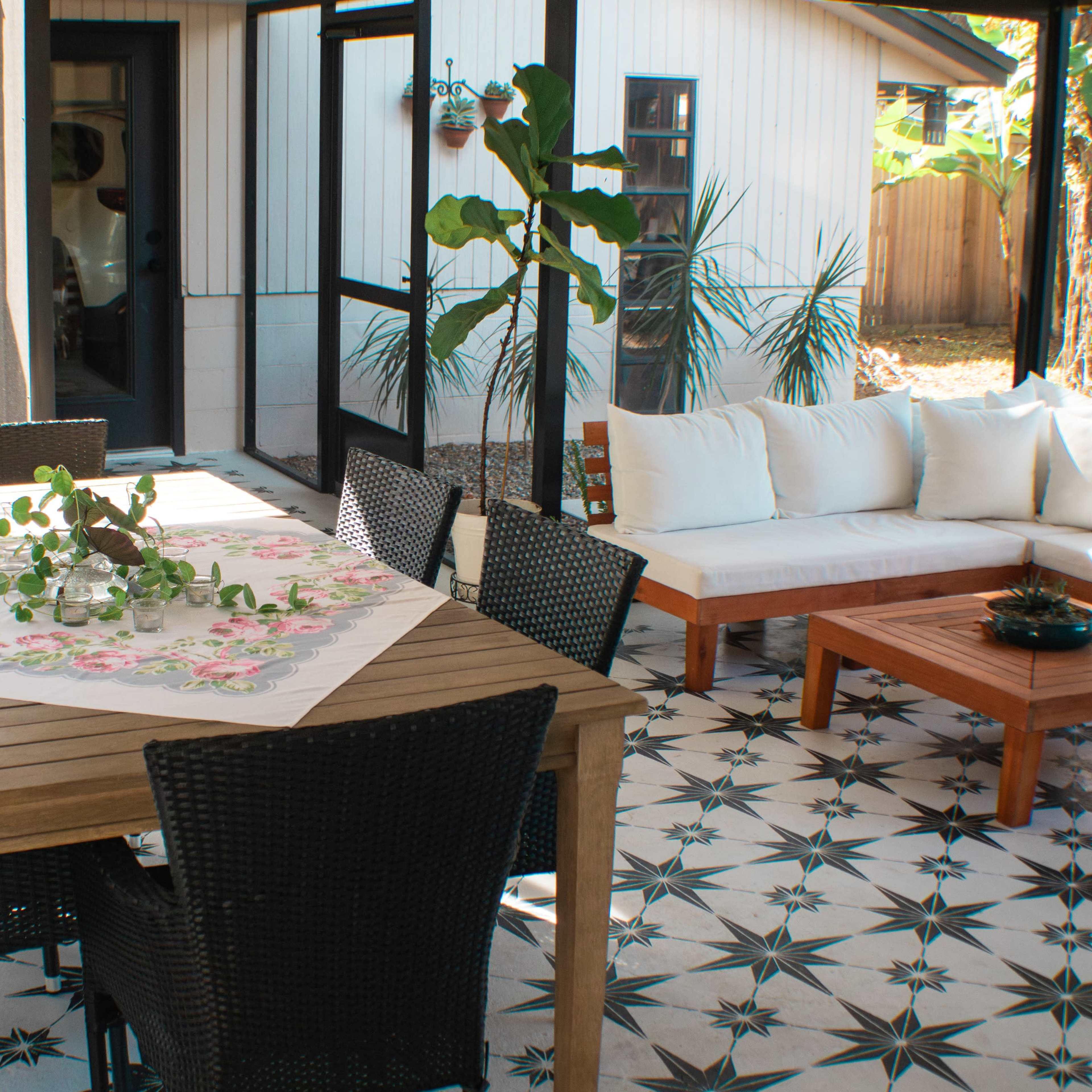 A modern outdoor patio with a wooden table surrounded by black chairs, a white sectional sofa, and patterned tile flooring.