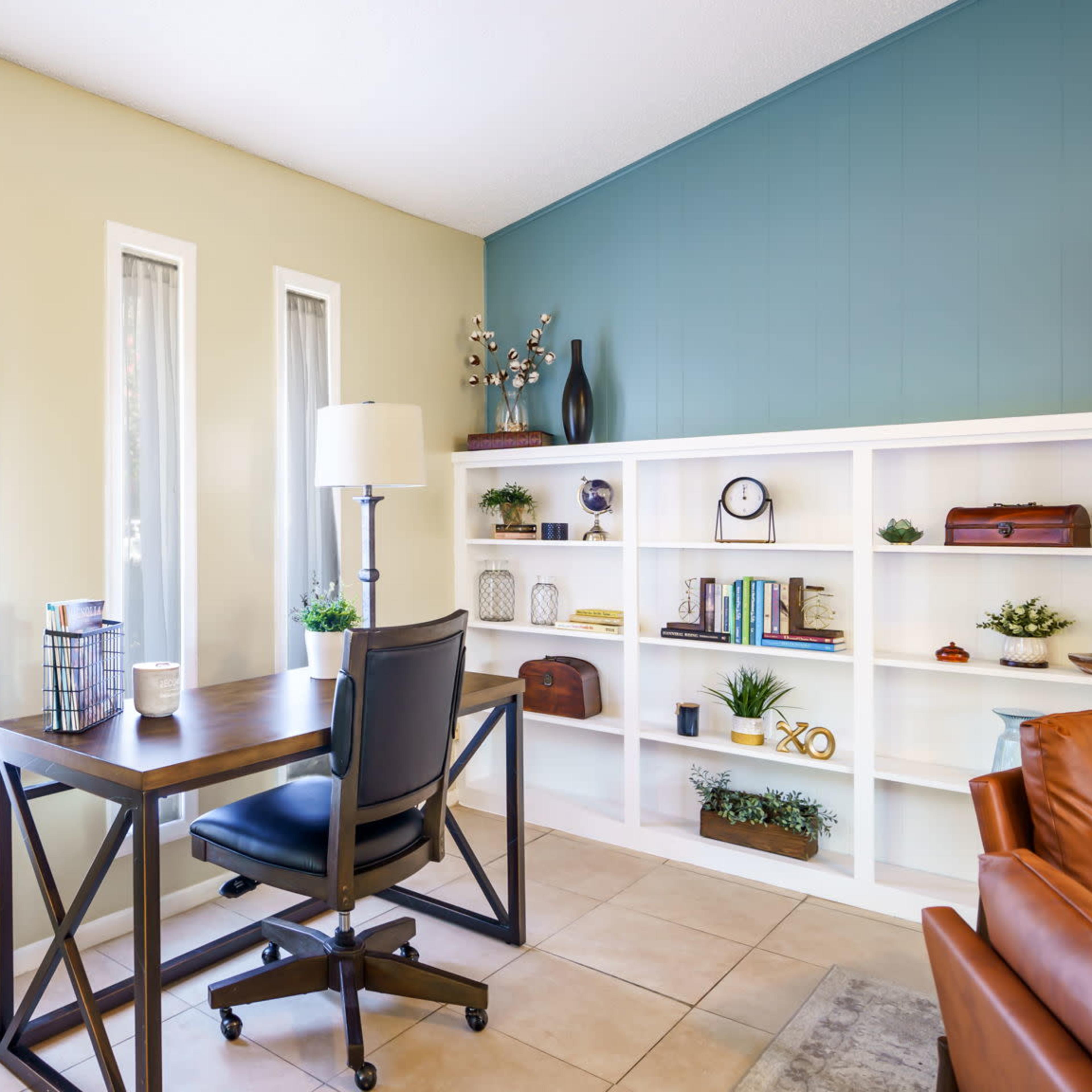 A home office features a wooden desk and black chair positioned against a wall with large windows, complemented by a bookshelf filled with decorative items and a potted plant.