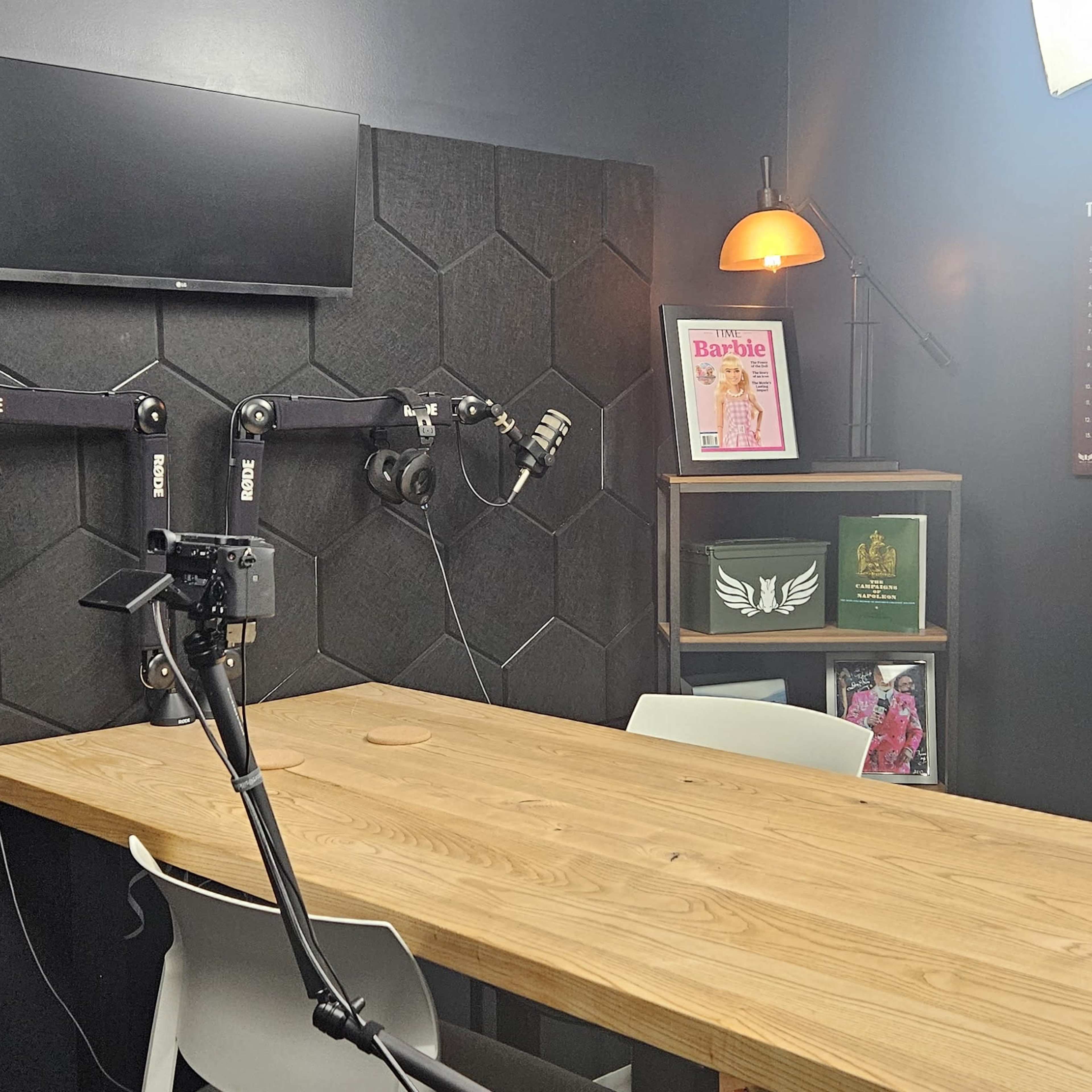 A podcast recording studio with a wooden table, multiple microphones mounted on the wall, and a television screen above, surrounded by soundproof panels and decorative shelves.