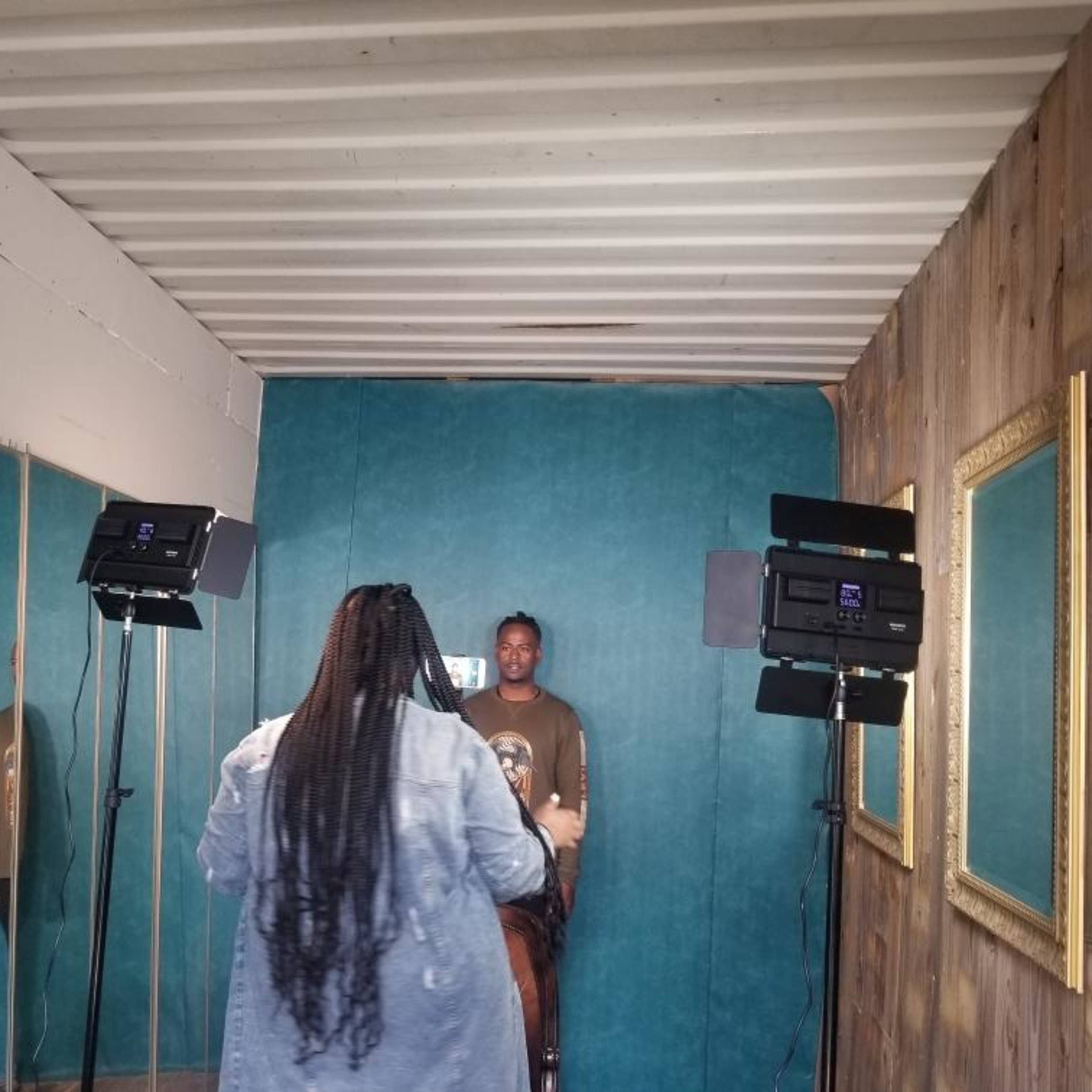 A person with long hair stands in front of a teal wall while a second person poses in front of a mirror under studio lights.