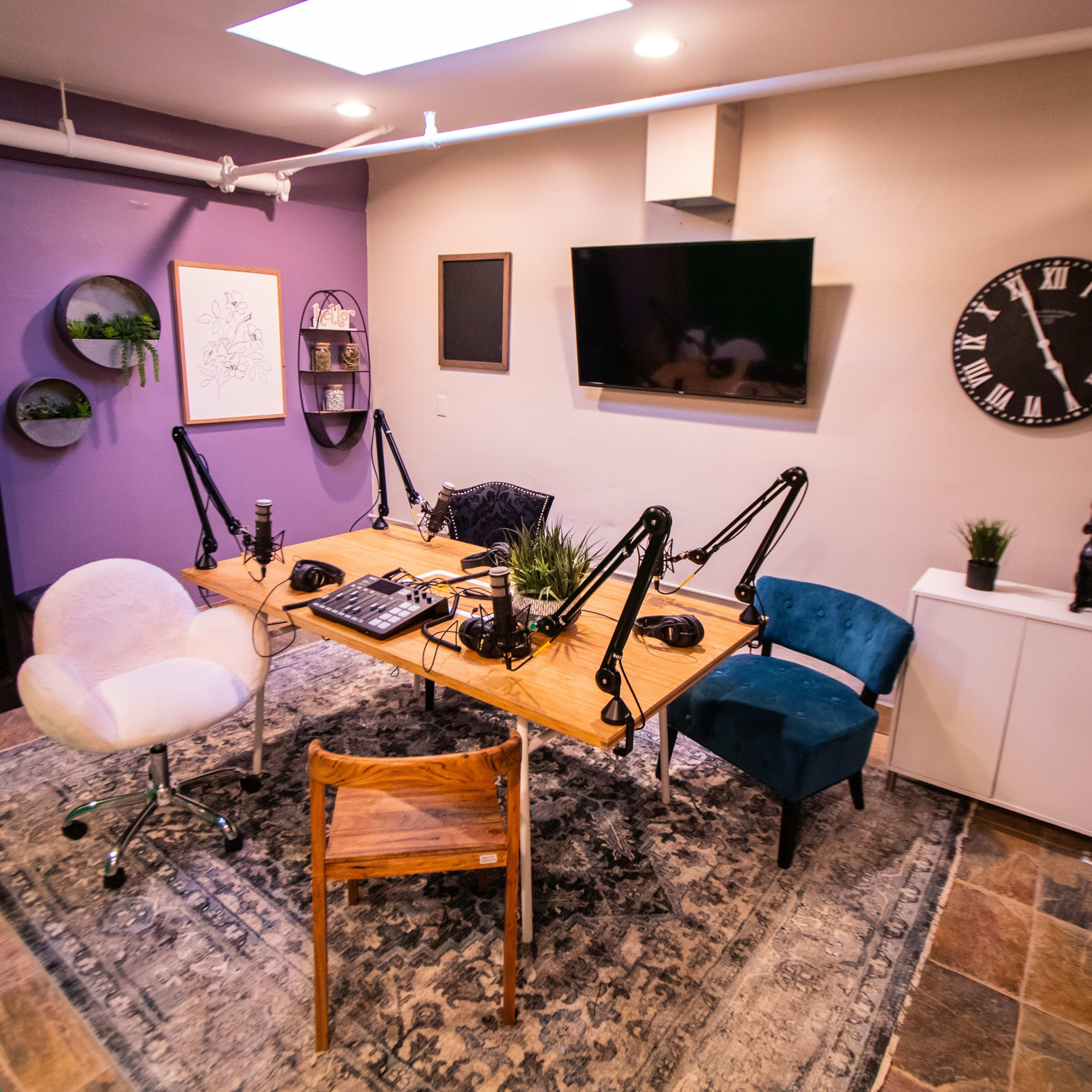 A podcast recording studio featuring a large wooden table with microphone setups, two chairs, and various decorative elements on the walls and furniture.