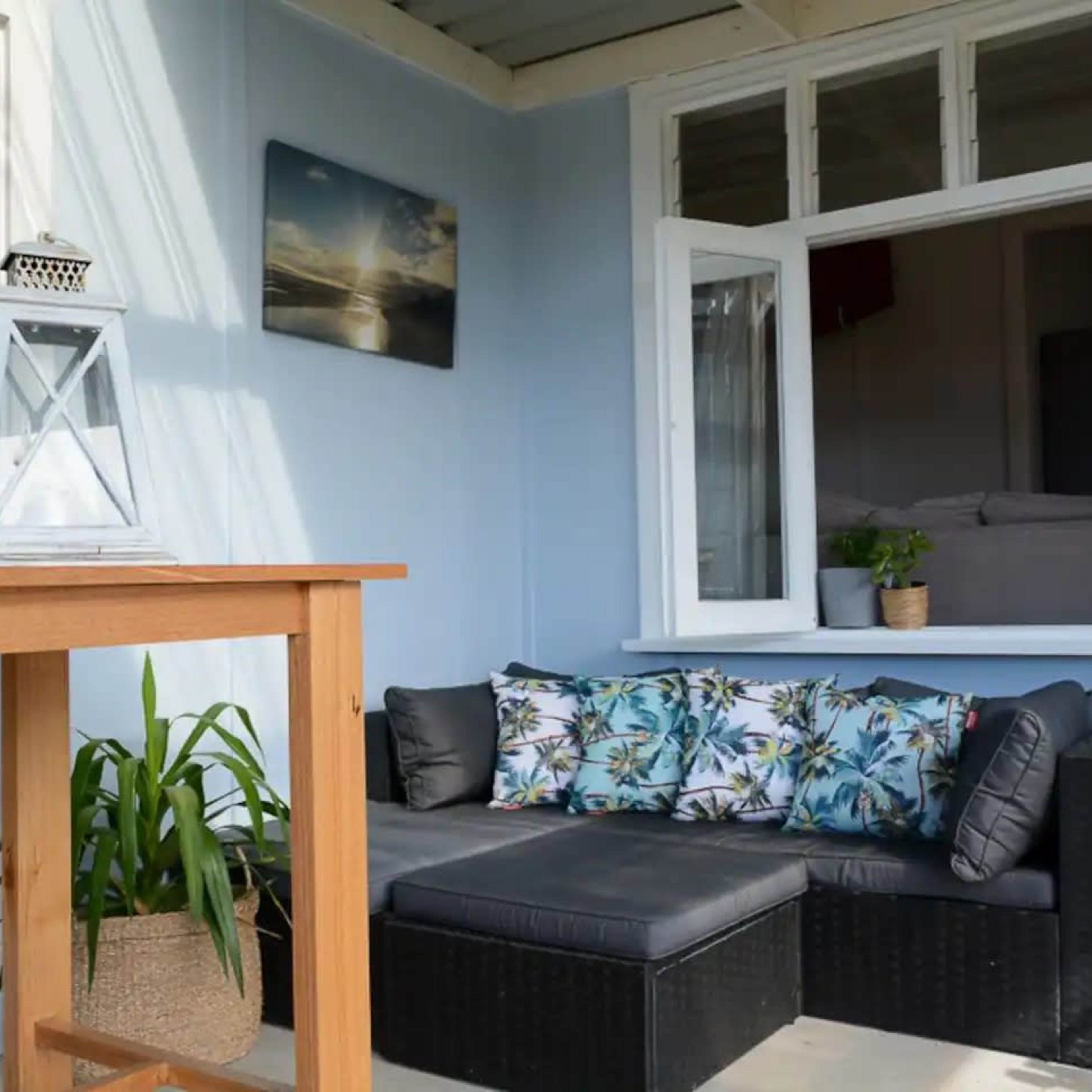 A small patio features a black wicker sofa with decorative cushions, a wooden table, a lantern, and an open window leading into a bedroom.