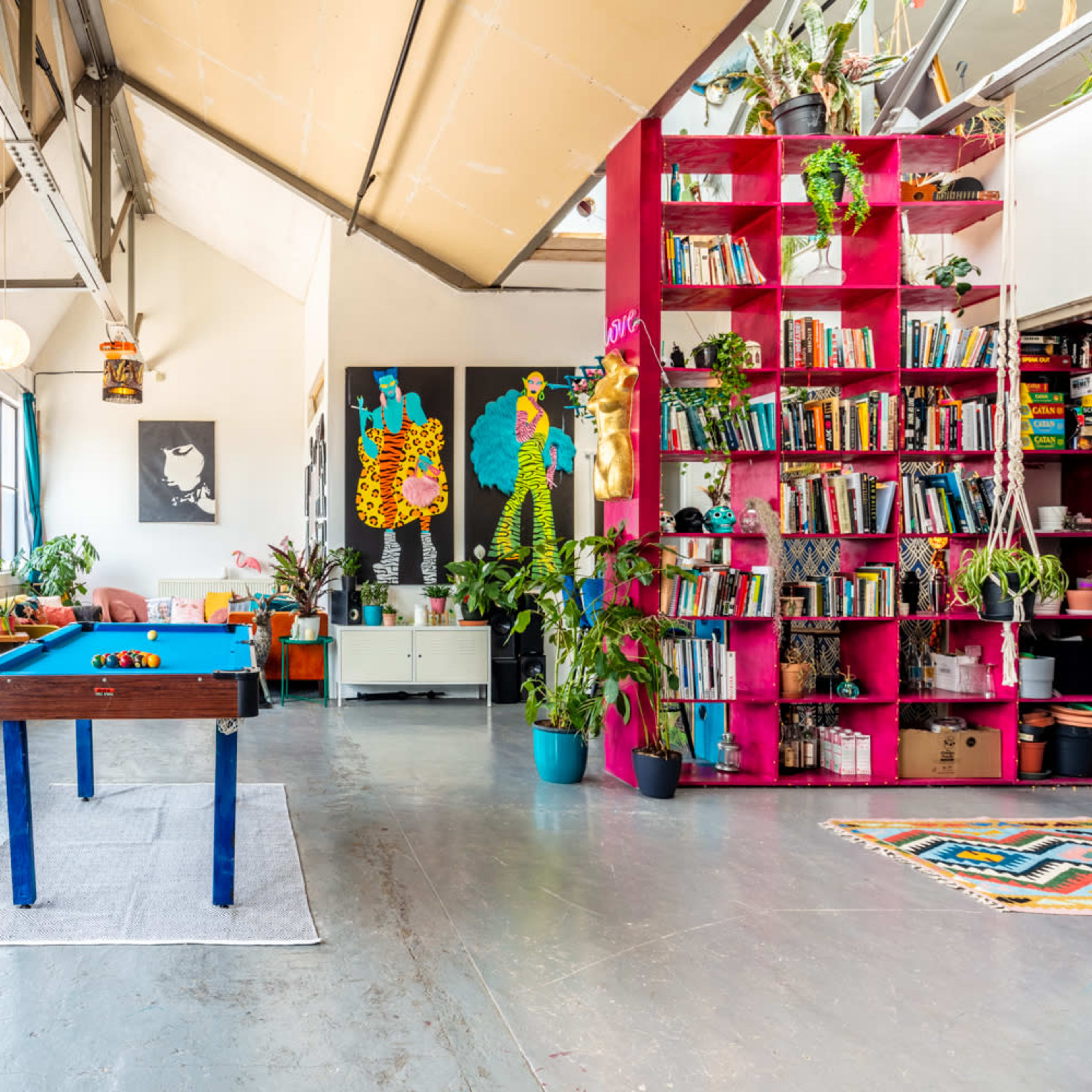 A bright, spacious loft with large windows, featuring a blue pool table, a bookshelf filled with books, and various indoor plants.