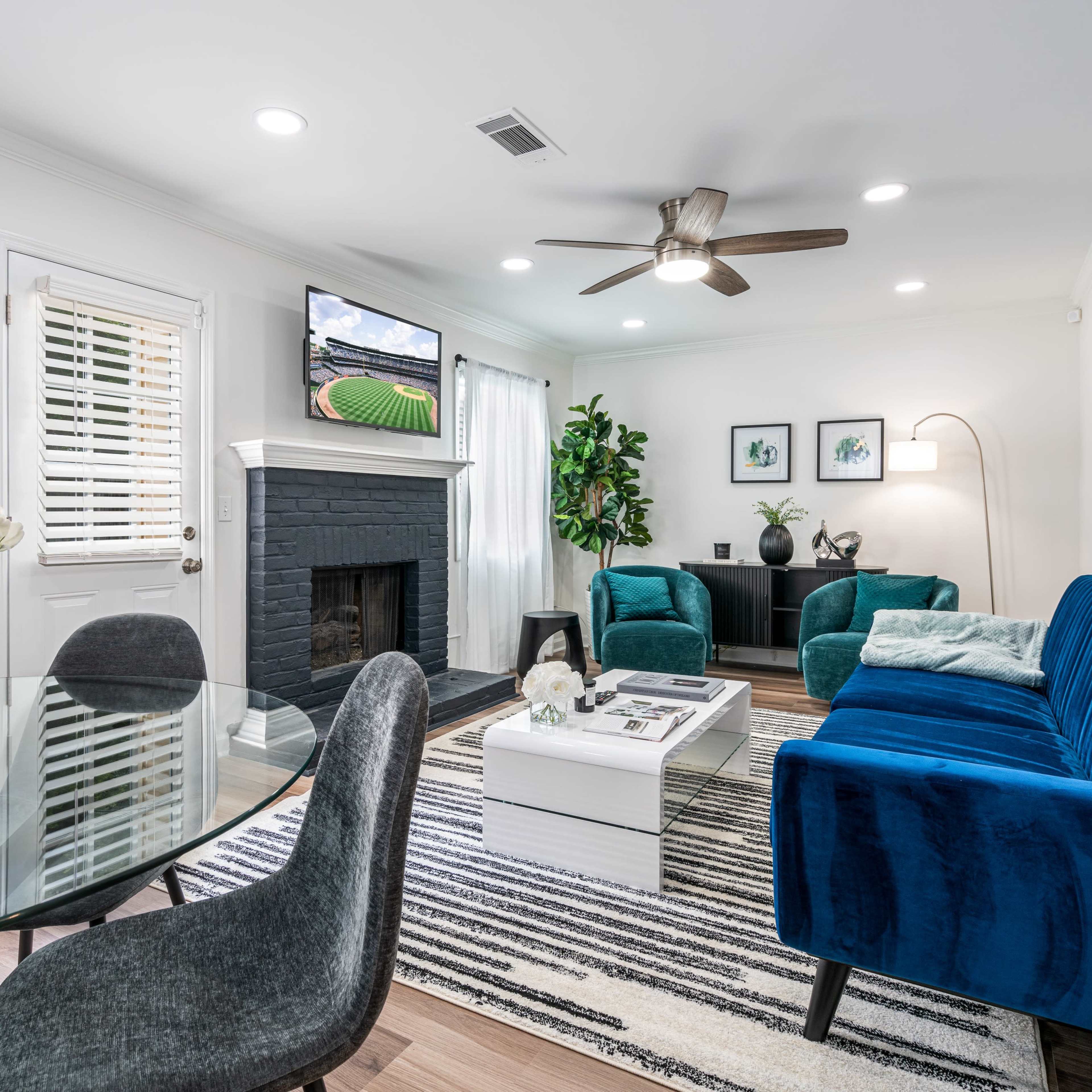 A modern living room with a blue velvet sofa, a glass dining table with floral decor, and a wall-mounted television above a black fireplace.