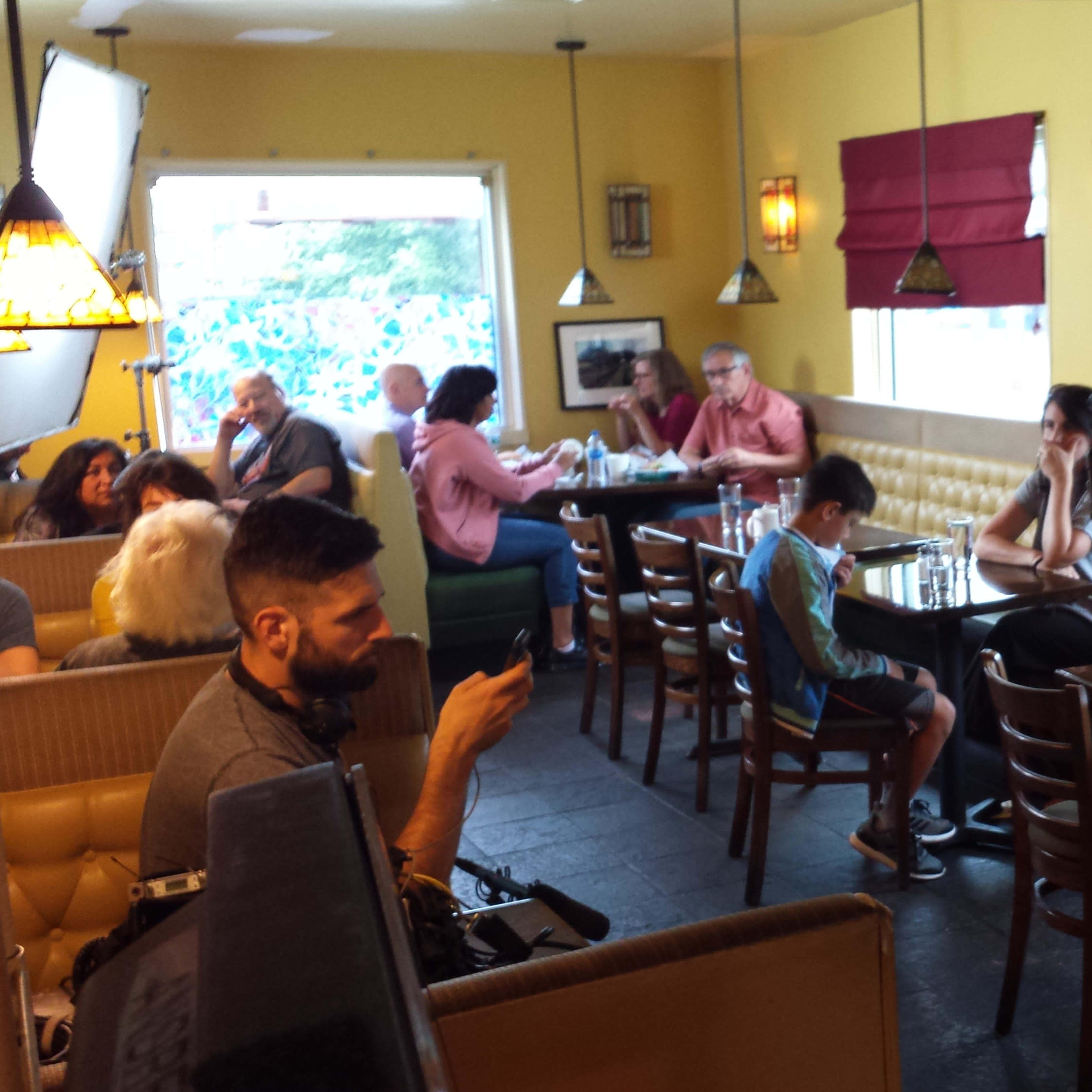 A film crew sets up cameras and equipment in a brightly lit restaurant, where diners are seated at various tables.
