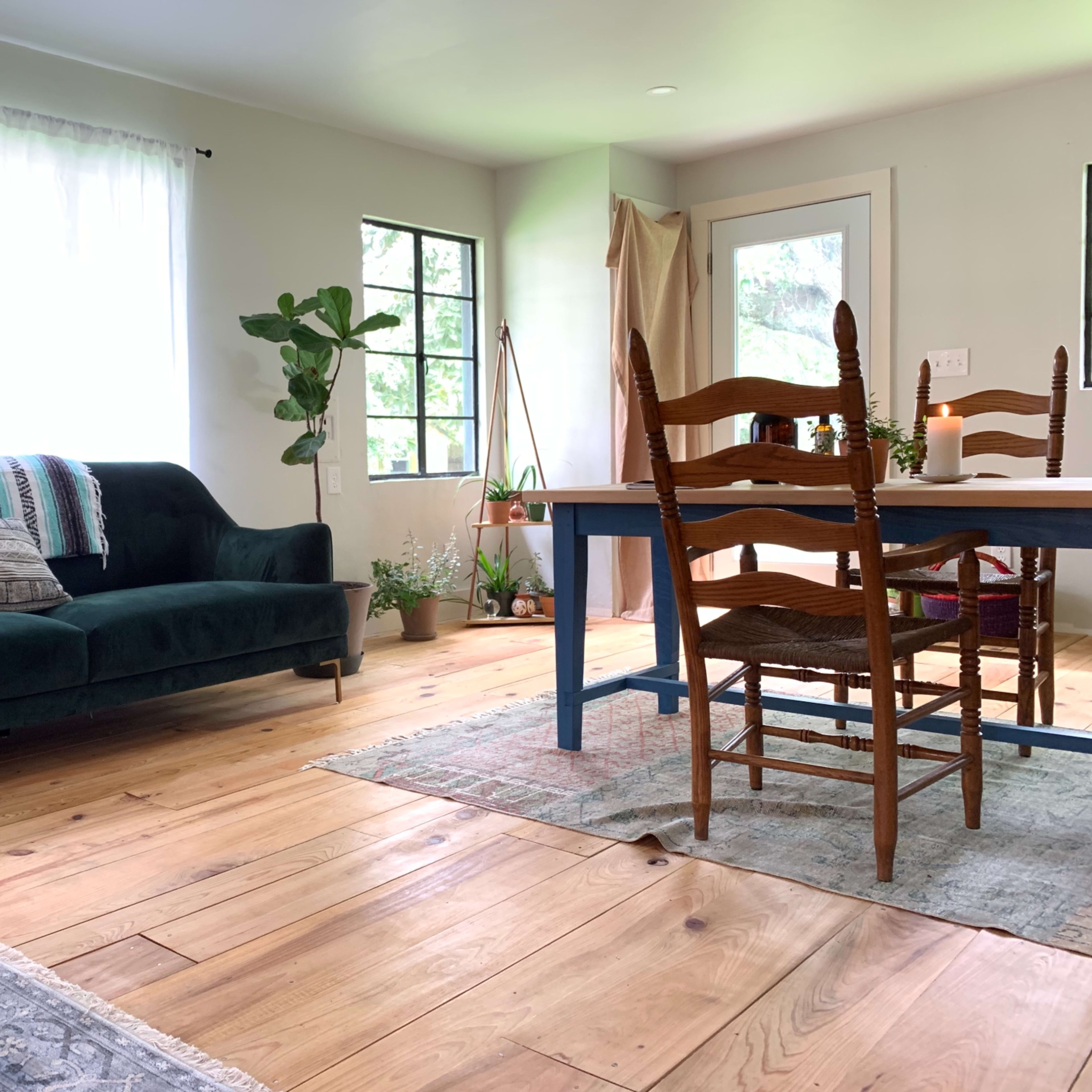 A living room with a green sofa, a wooden dining table with chairs, and large windows allowing natural light to enter.