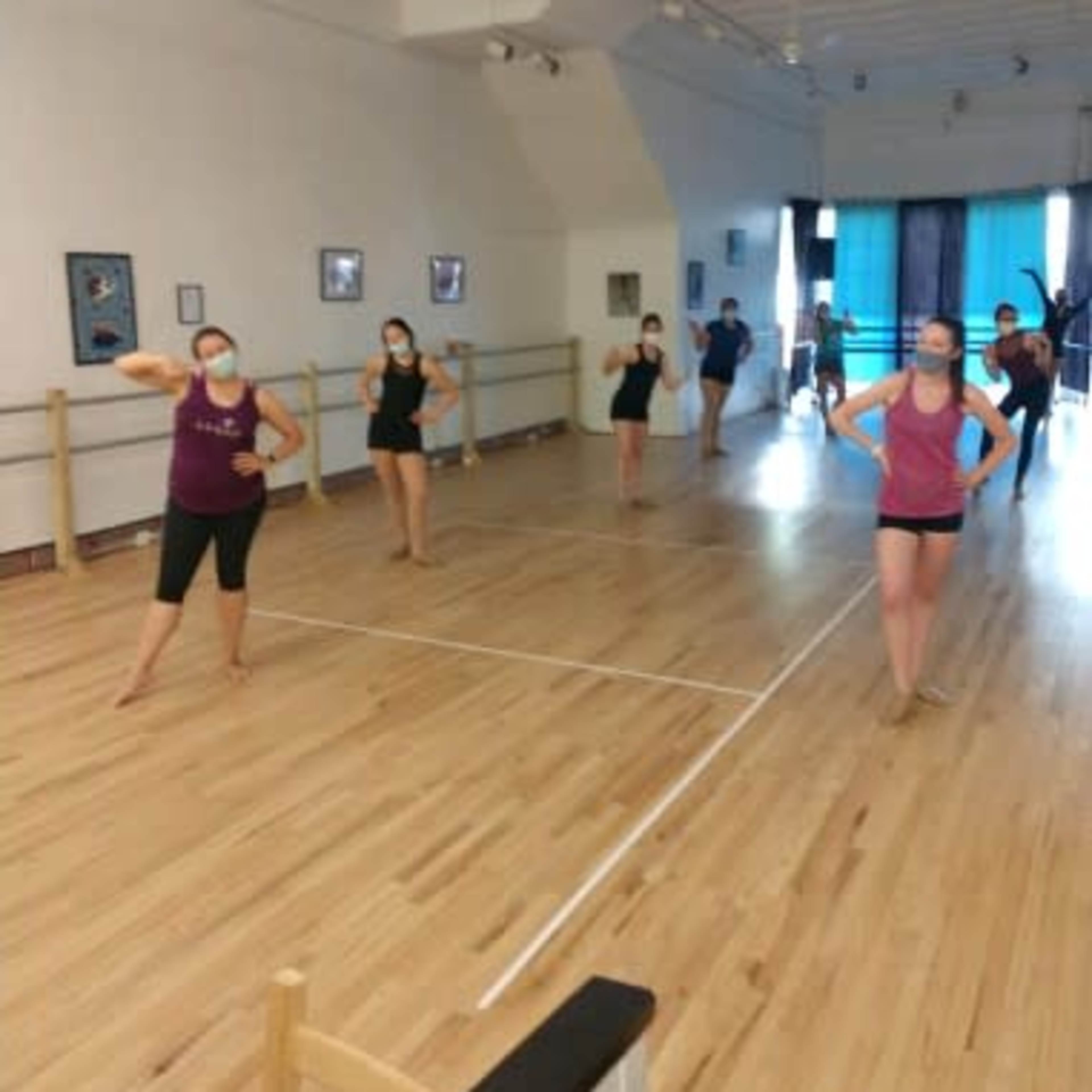 A group of dancers in active pose rehearses in a wooden-floored studio with mirrors and a ballet barre.