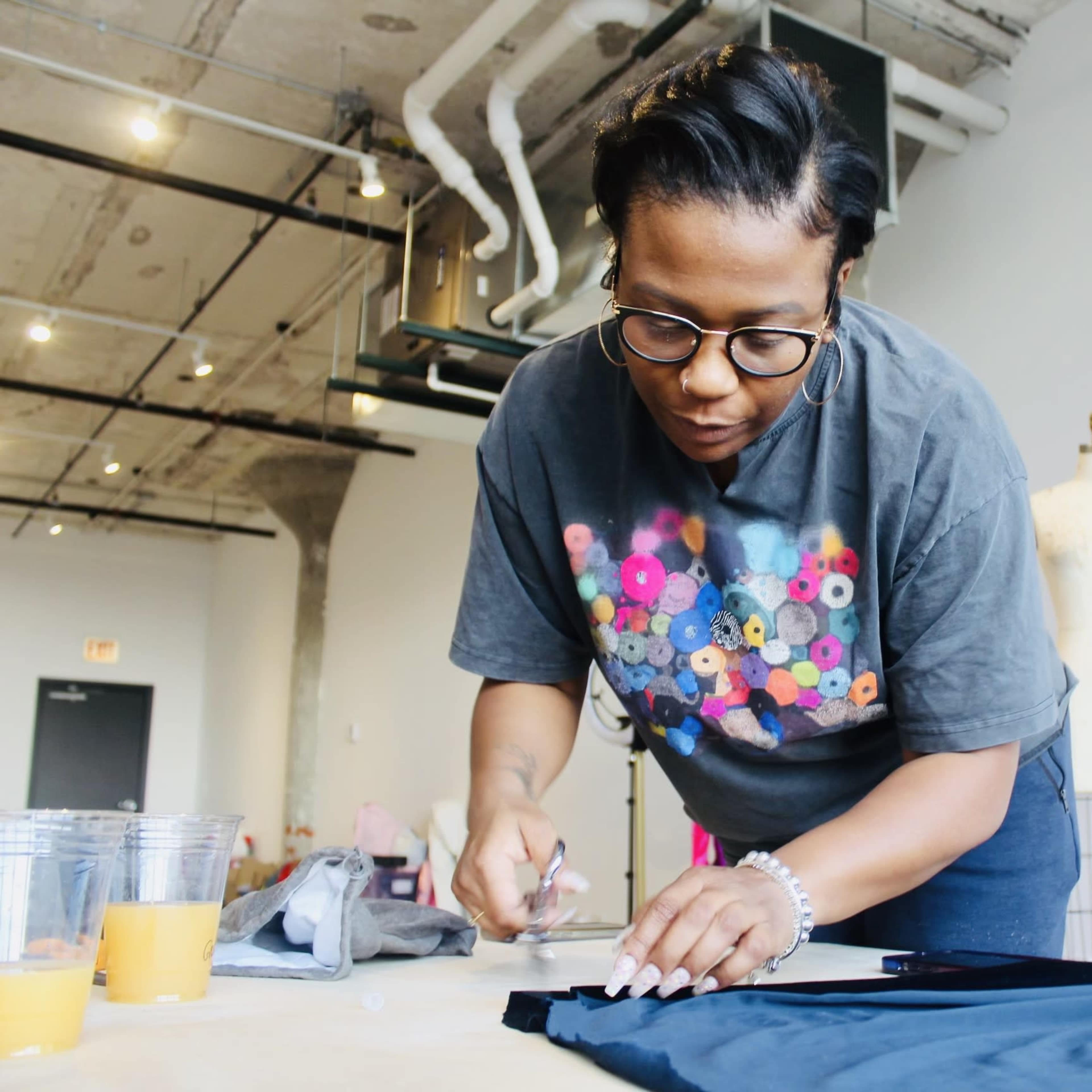 A woman in a colorful t-shirt is cutting fabric on a table in a workshop space.