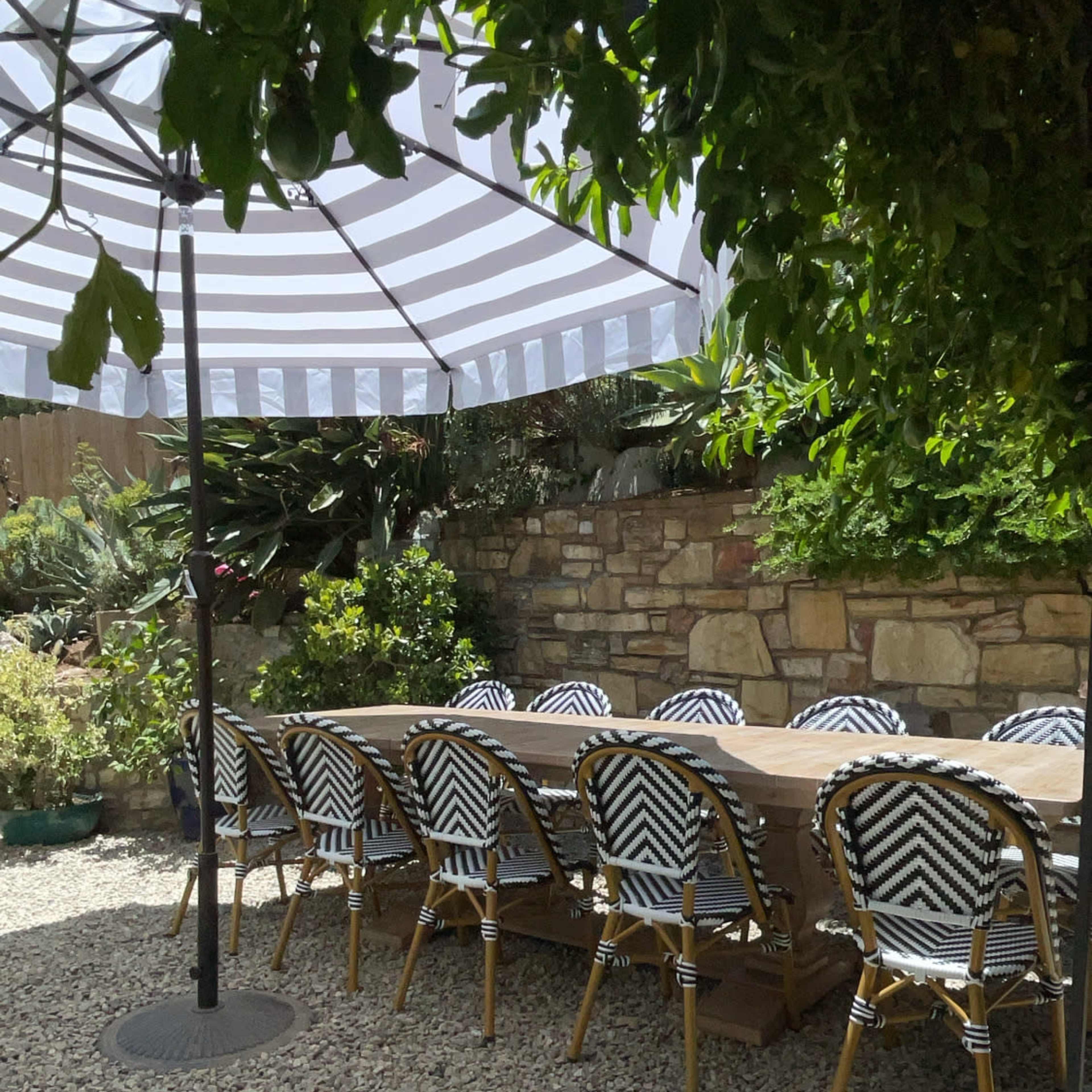 A large, rectangular wooden table is surrounded by patterned chairs beneath a striped umbrella in a shaded outdoor area with gravel.