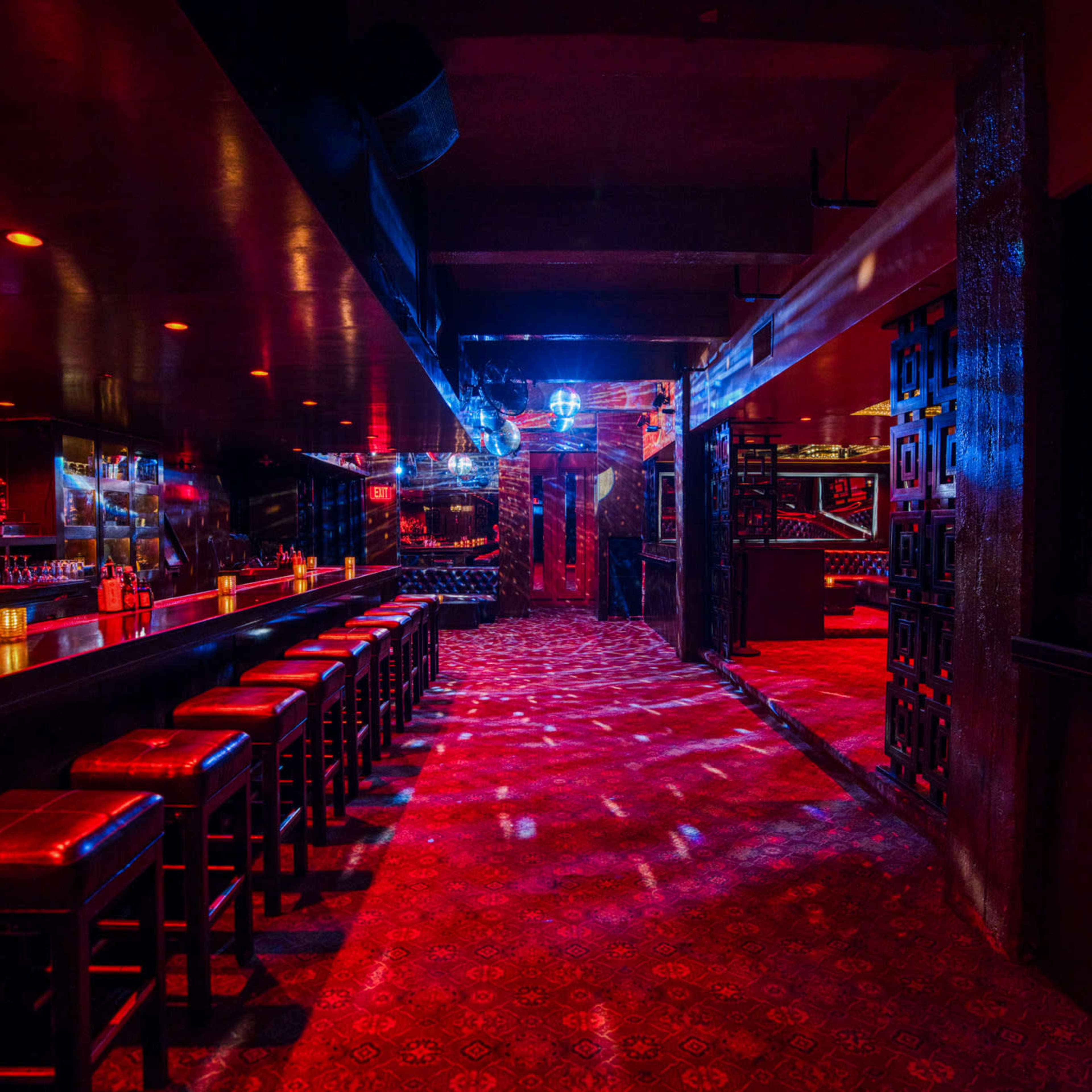 The dimly lit interior of a bar features a long counter with high stools and patterned carpeting, illuminated by colorful lights.