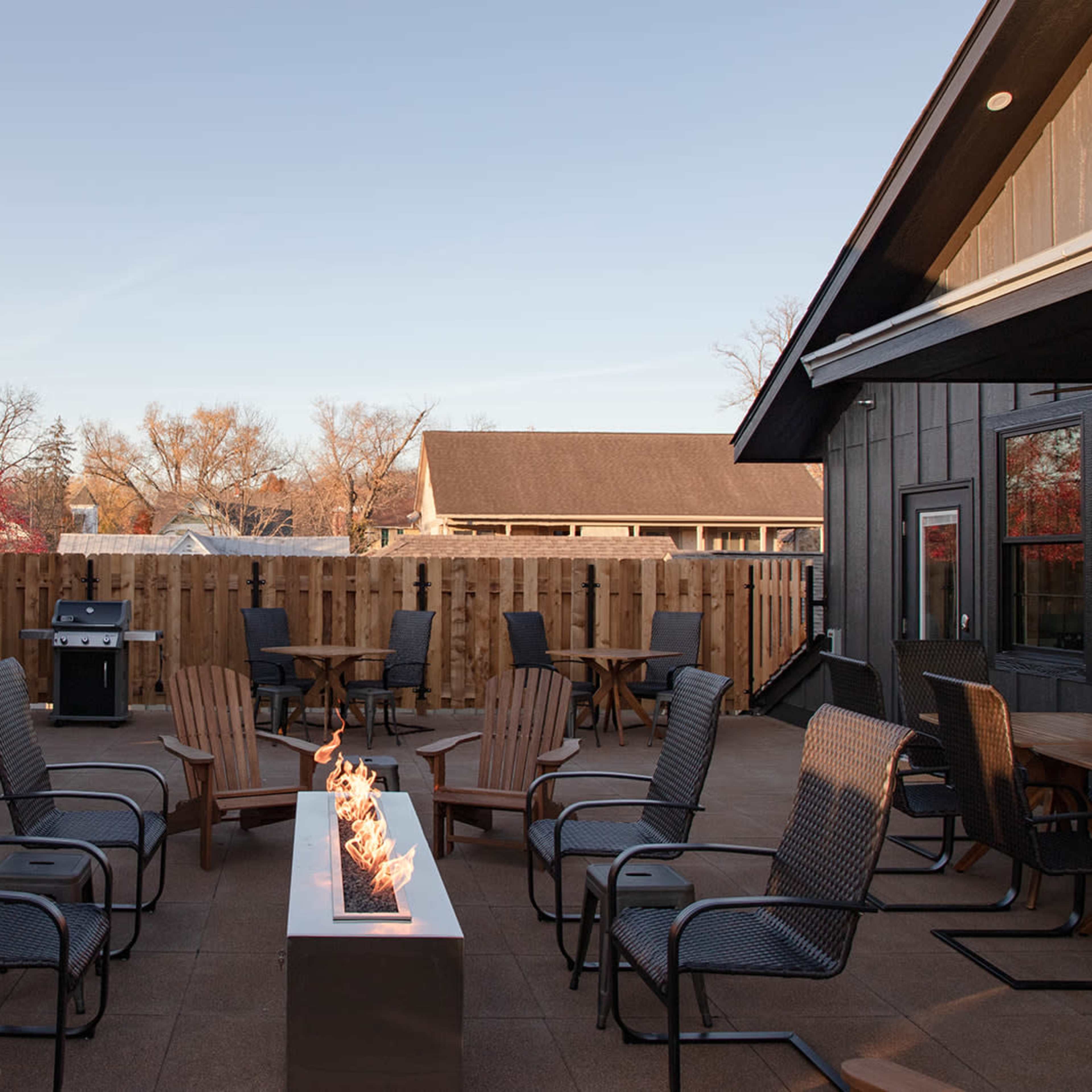 A patio area features several chairs arranged around a fire pit, with a grill and wooden fencing in the background.