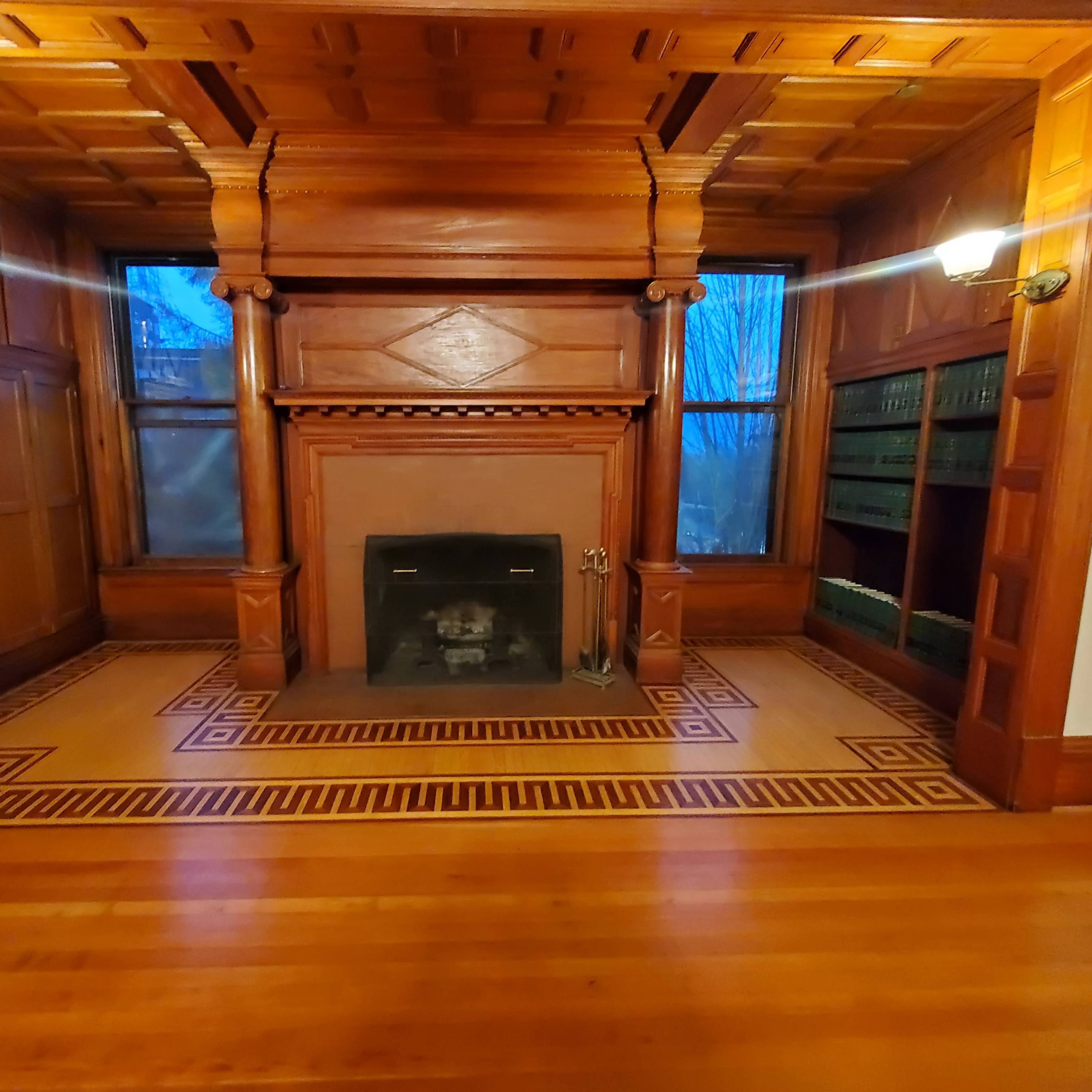 A wooden-paneled room with a fireplace at the center, flanked by wall-mounted lamps and a patterned floor.