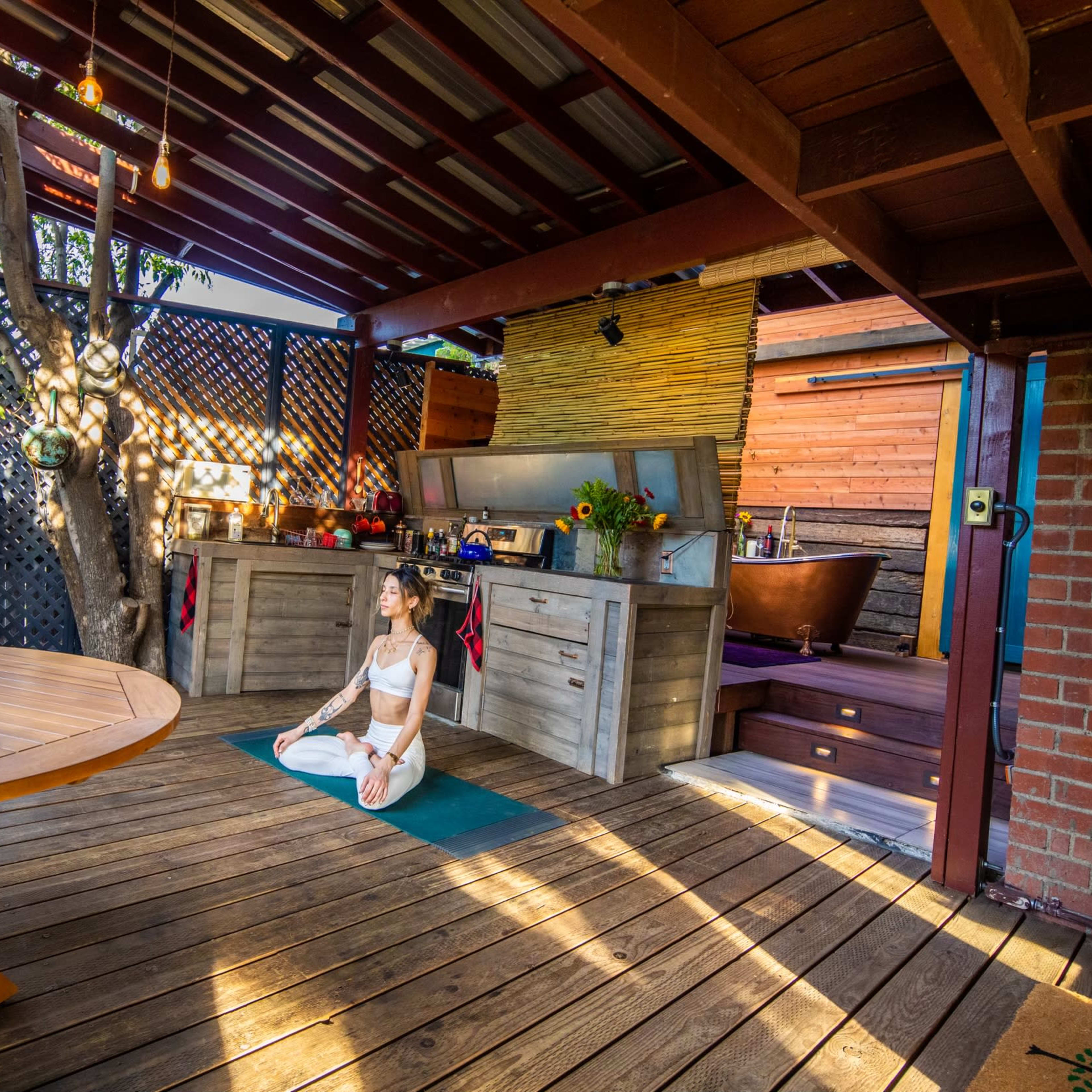 A woman practices yoga on a mat in a wooden deck area with an open kitchen and plants surrounding her.
