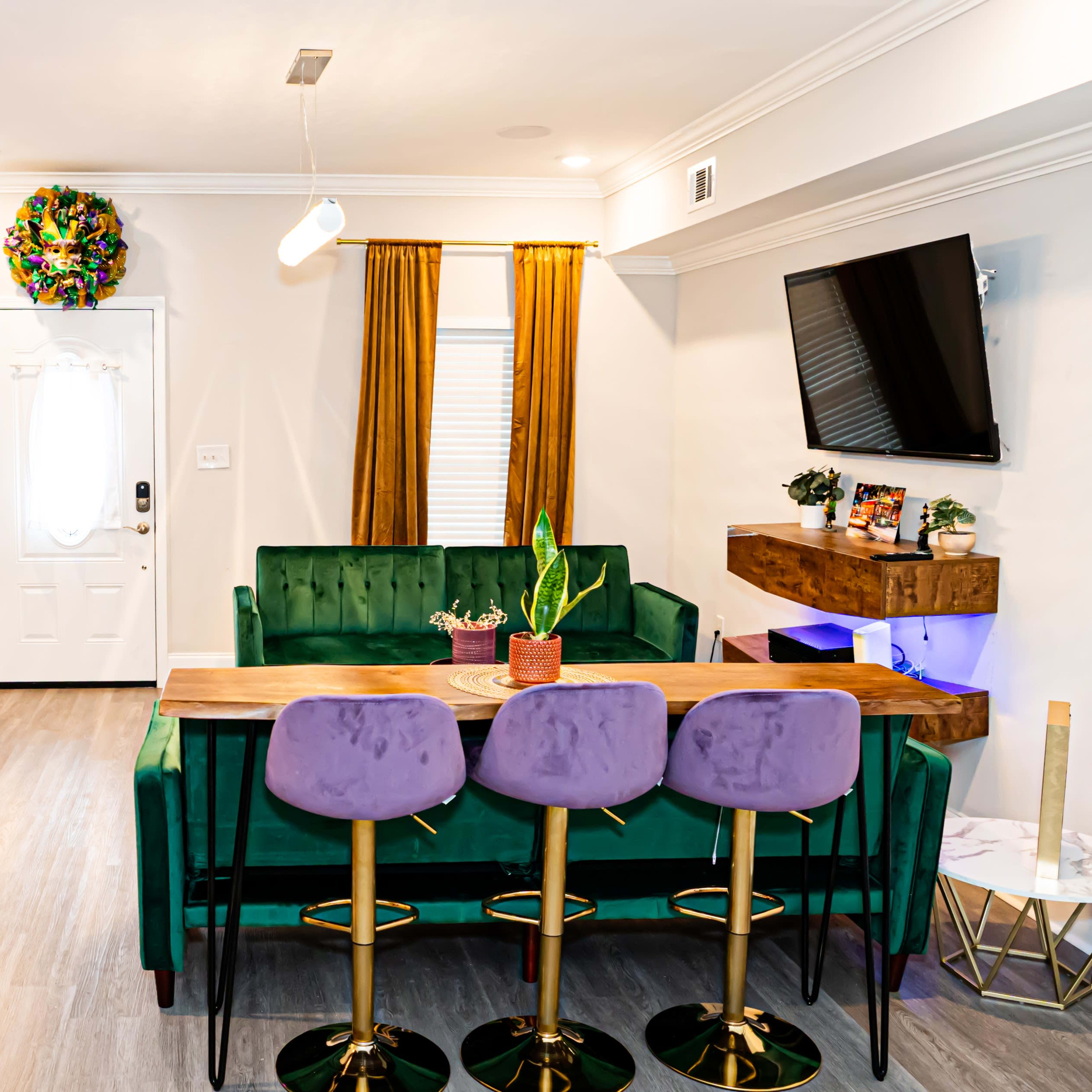 A modern living space with a green sofa, a wooden dining table surrounded by pink bar stools, a plant, and a decorative green wall with mirrors.