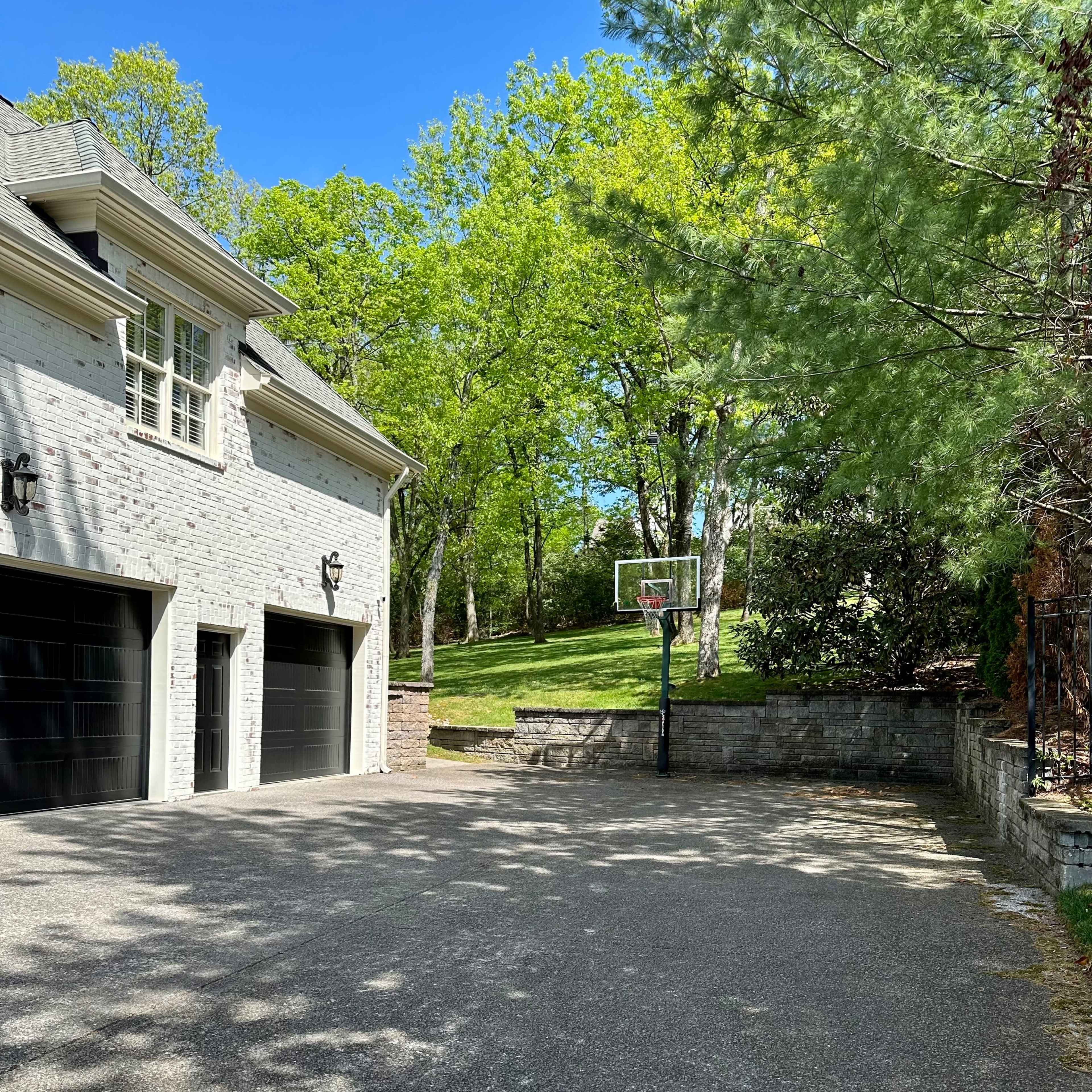 A driveway beside a house, featuring a basketball hoop and a grassy area in the background.