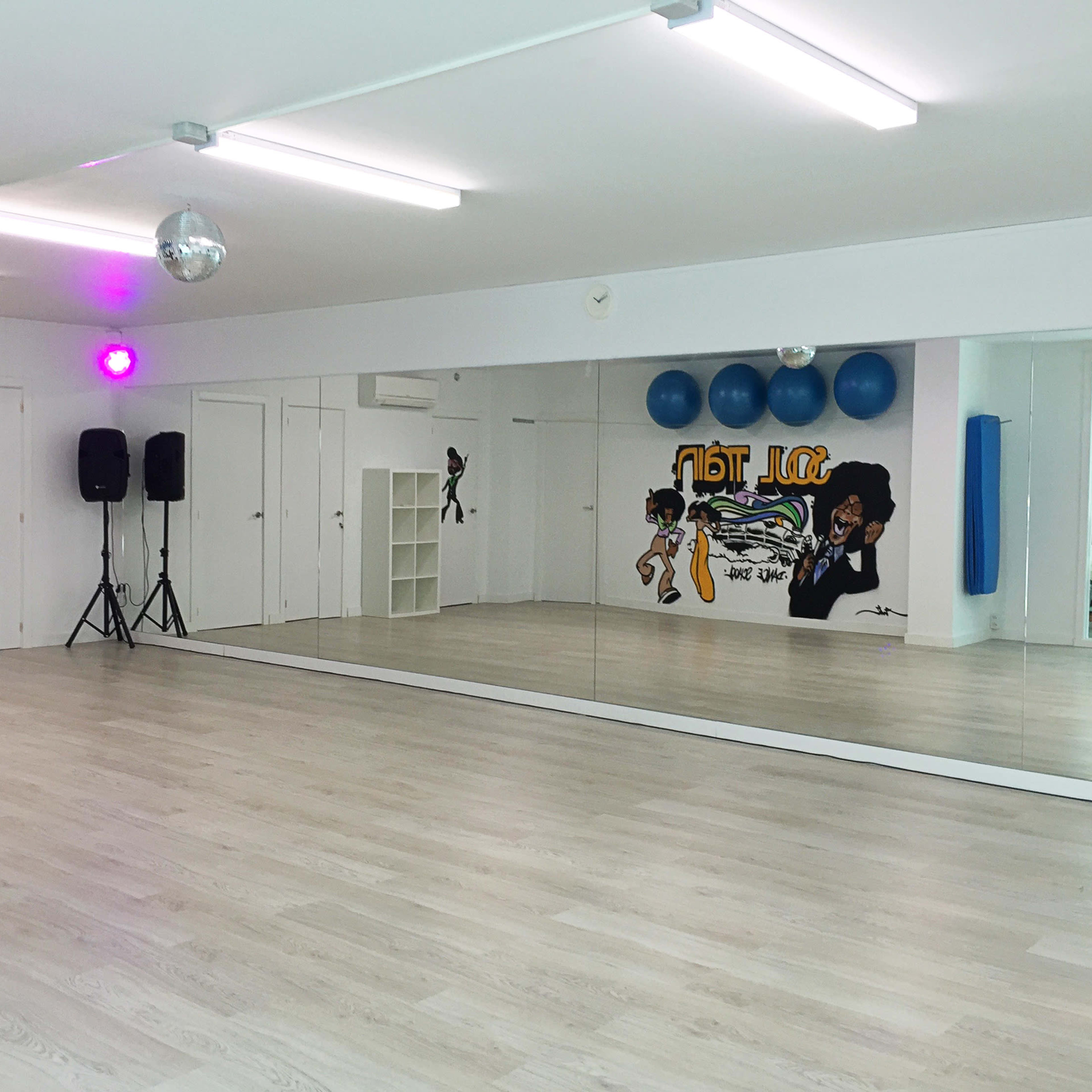 A dance studio with a mirrored wall, wooden flooring, and a colorful mural alongside blue exercise balls.