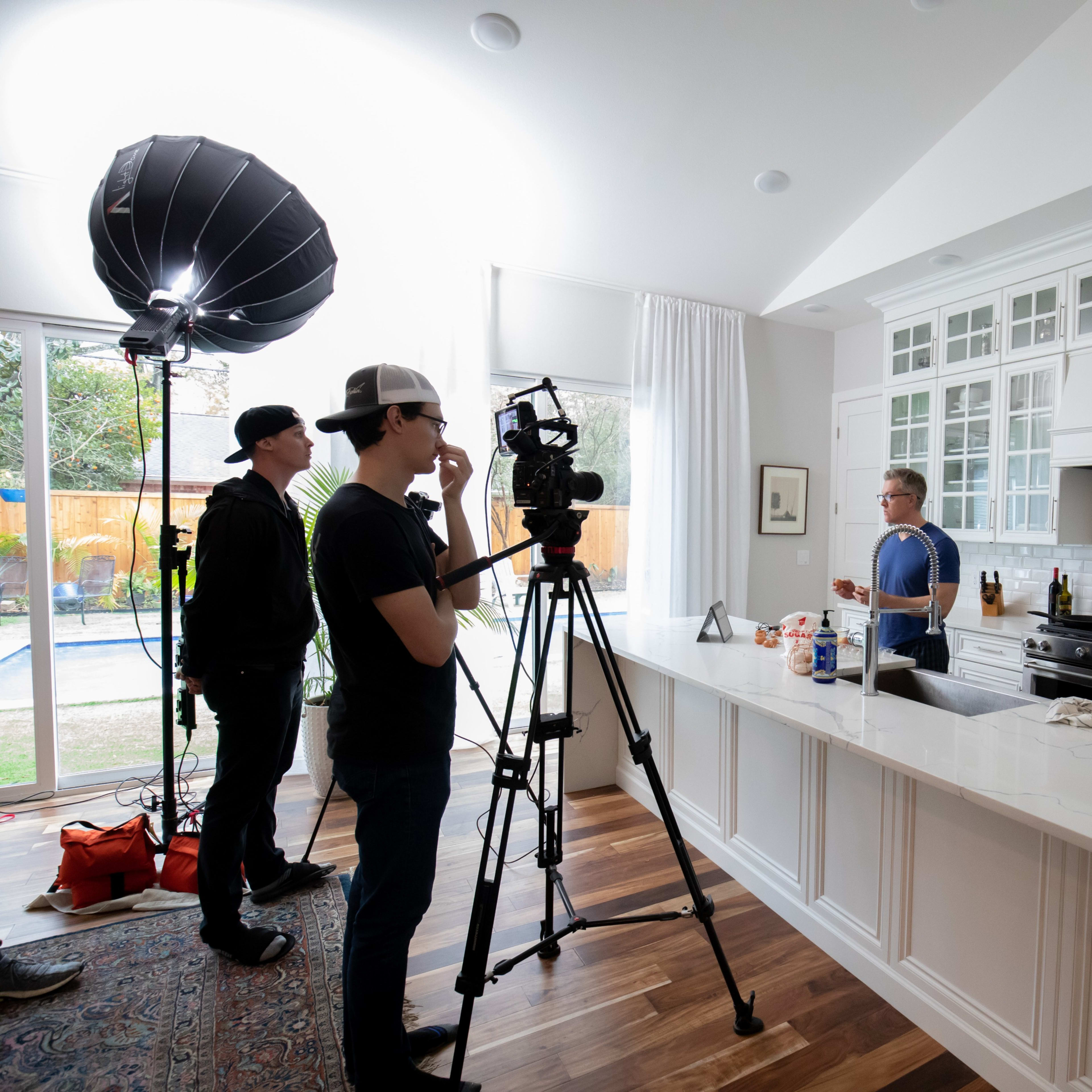 A film crew sets up to record a cooking segment in a modern kitchen with white cabinetry and a large island.