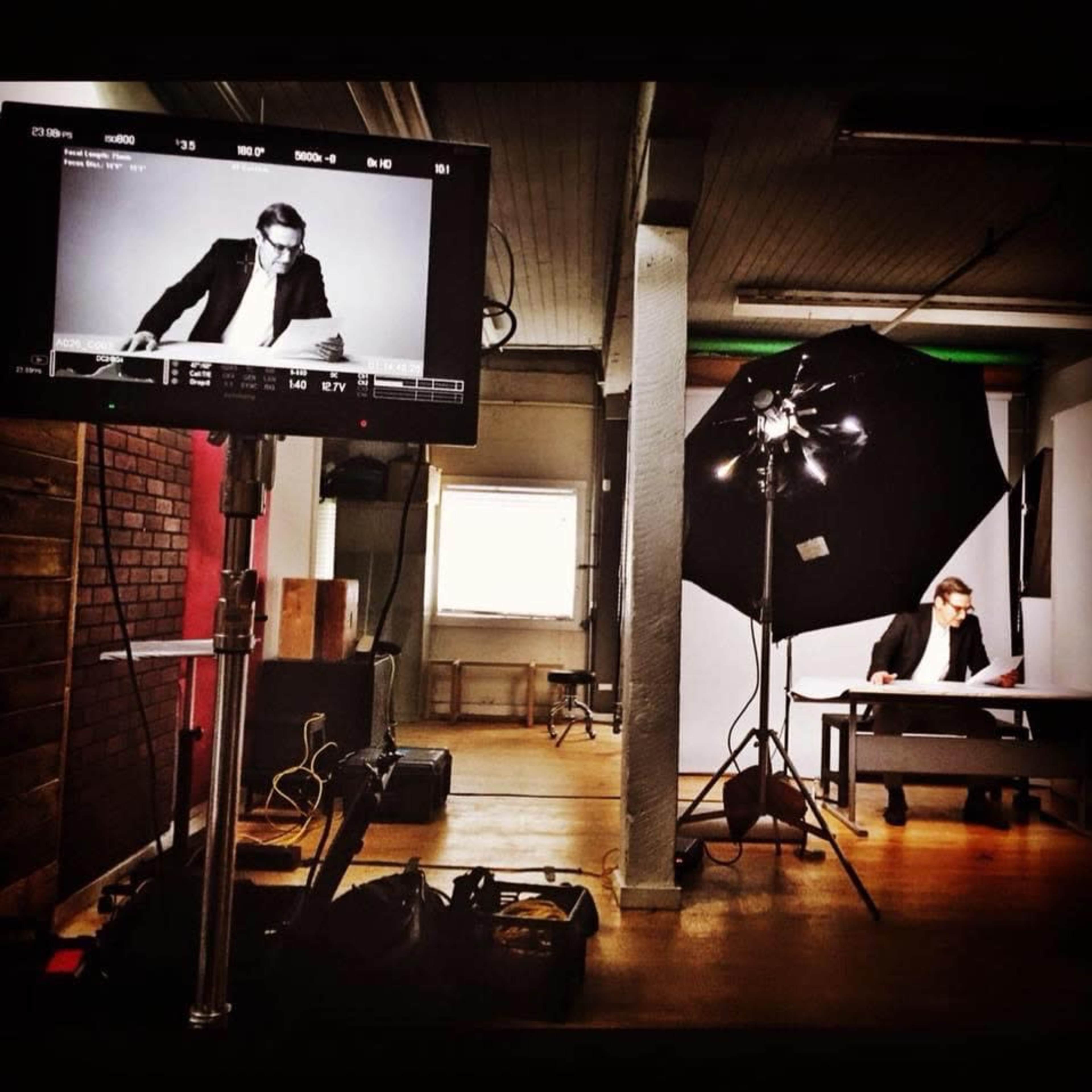 A photography studio setup shows a man in a suit sitting at a table, with a camera monitor and a large softbox light nearby.