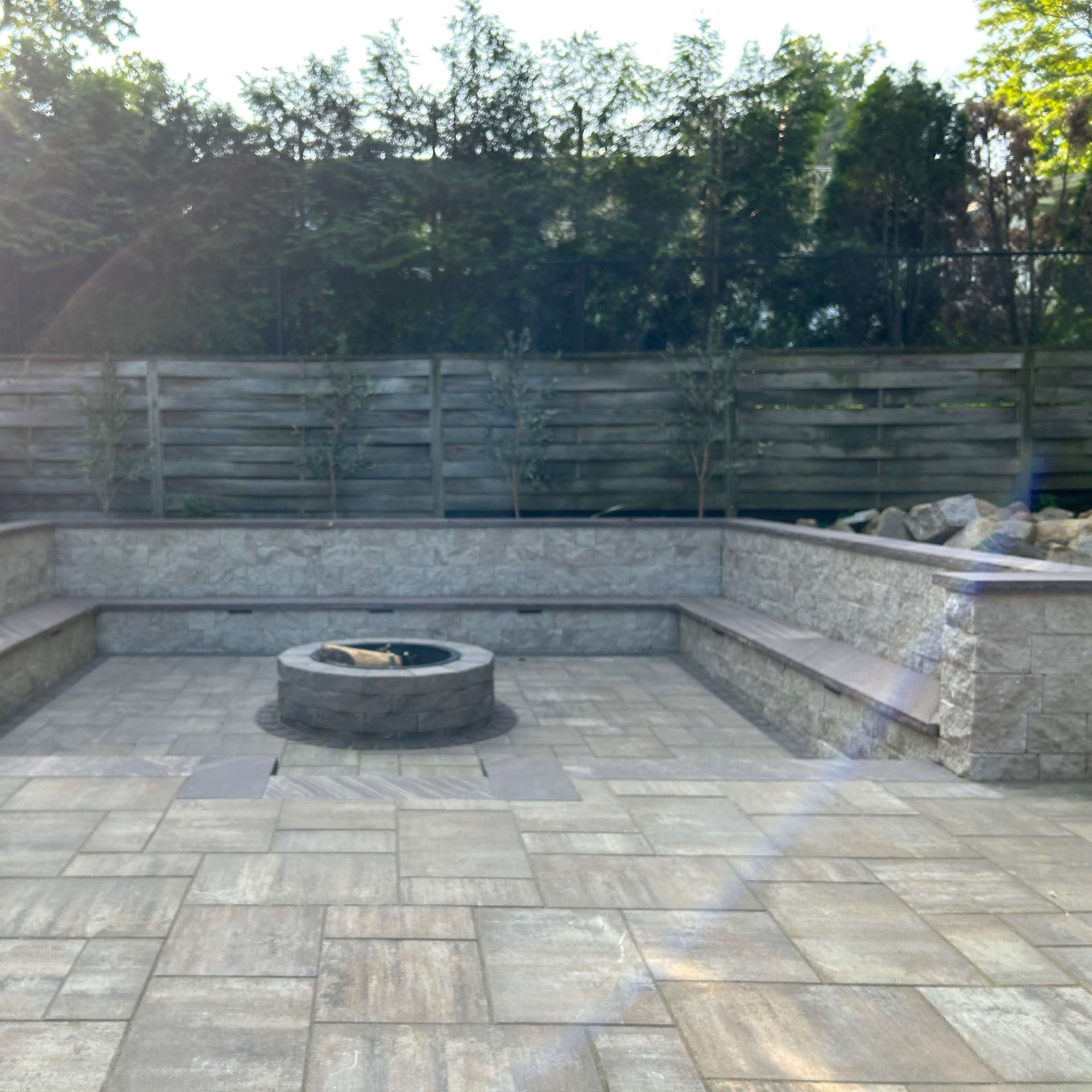 A stone patio with a central fire pit surrounded by built-in seating, enclosed by a wooden fence and greenery.