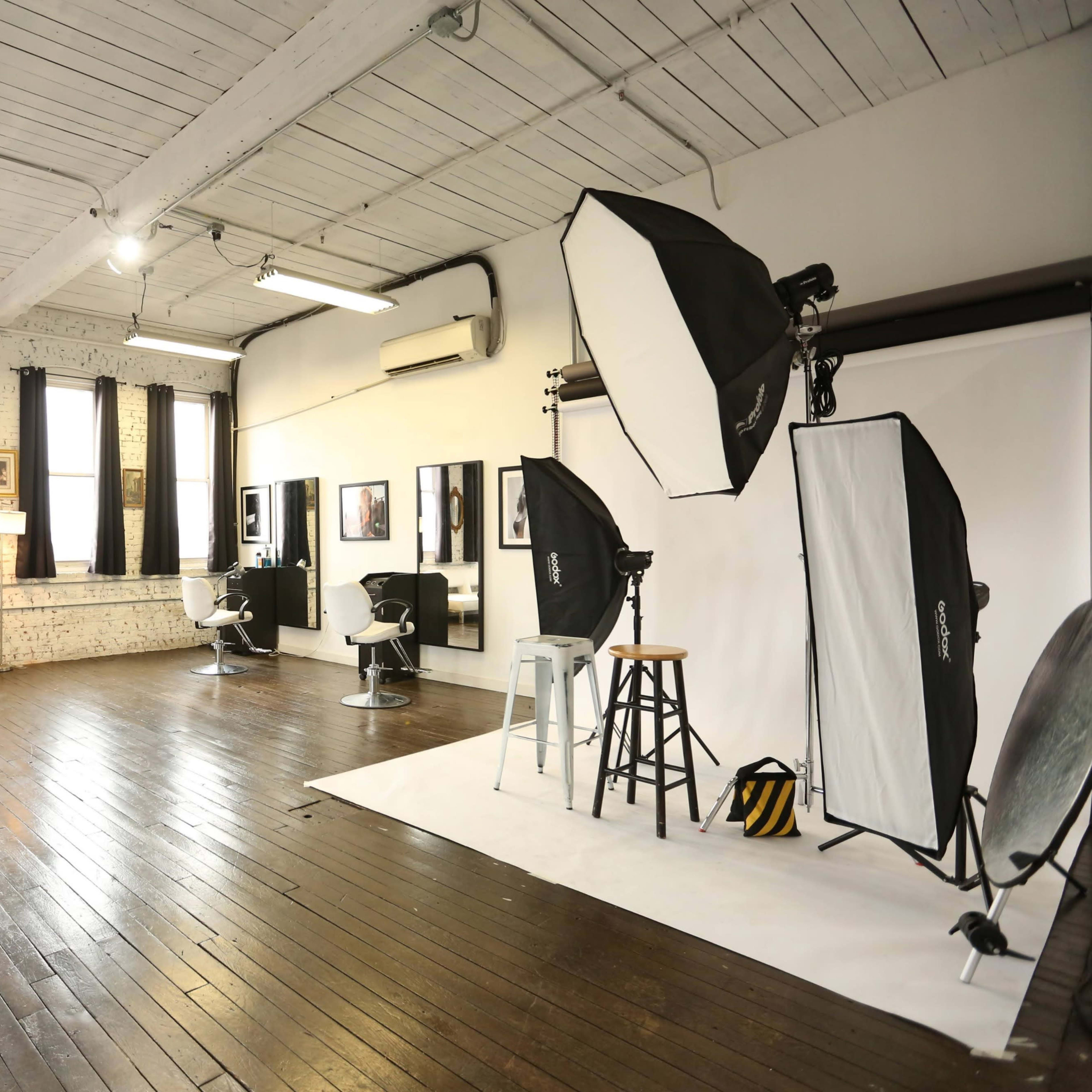 A spacious photography studio with wooden floors, large windows, and various lighting equipment arranged around a backdrop.