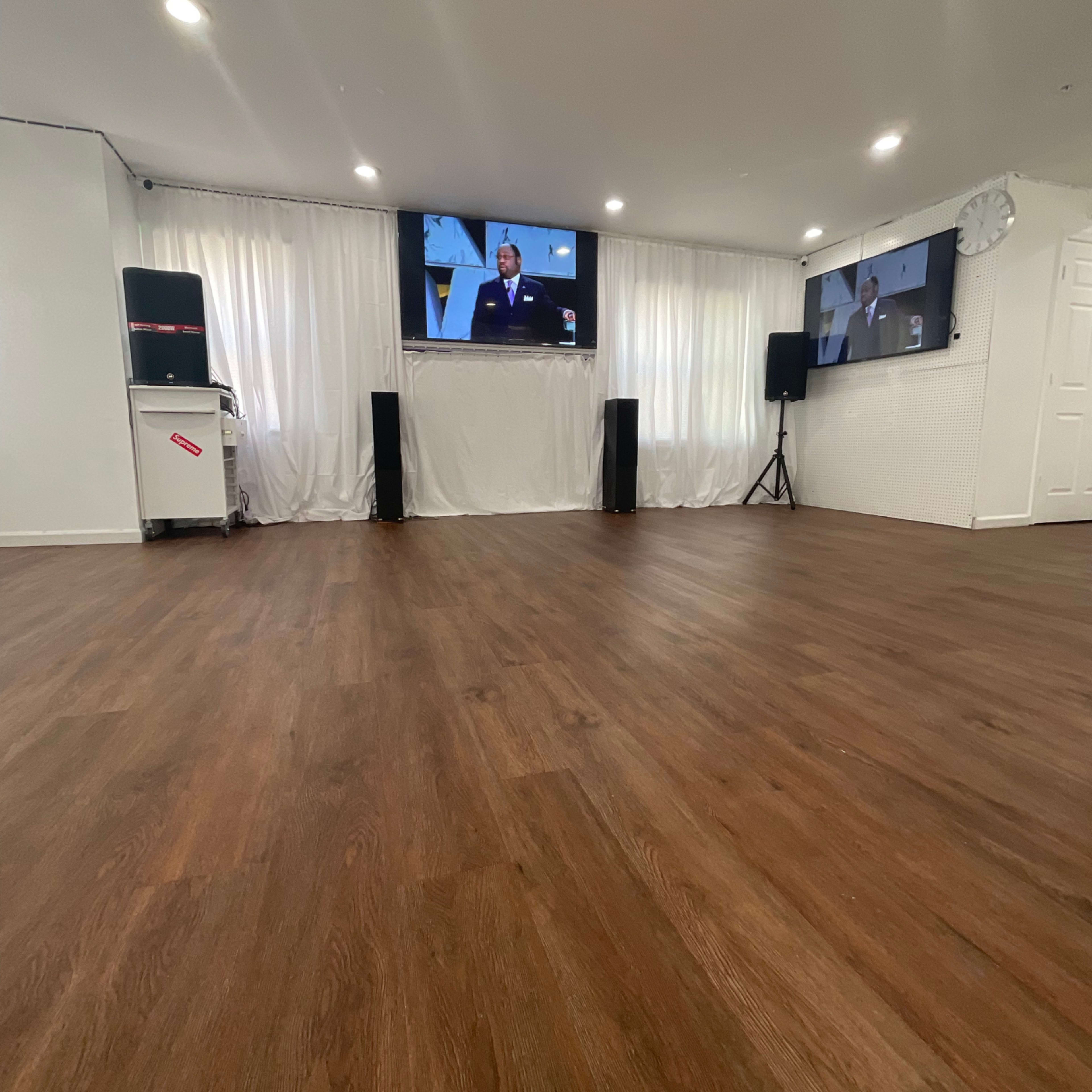 A spacious room with wooden flooring, two large television screens mounted on the walls, and sound speakers positioned at the sides.