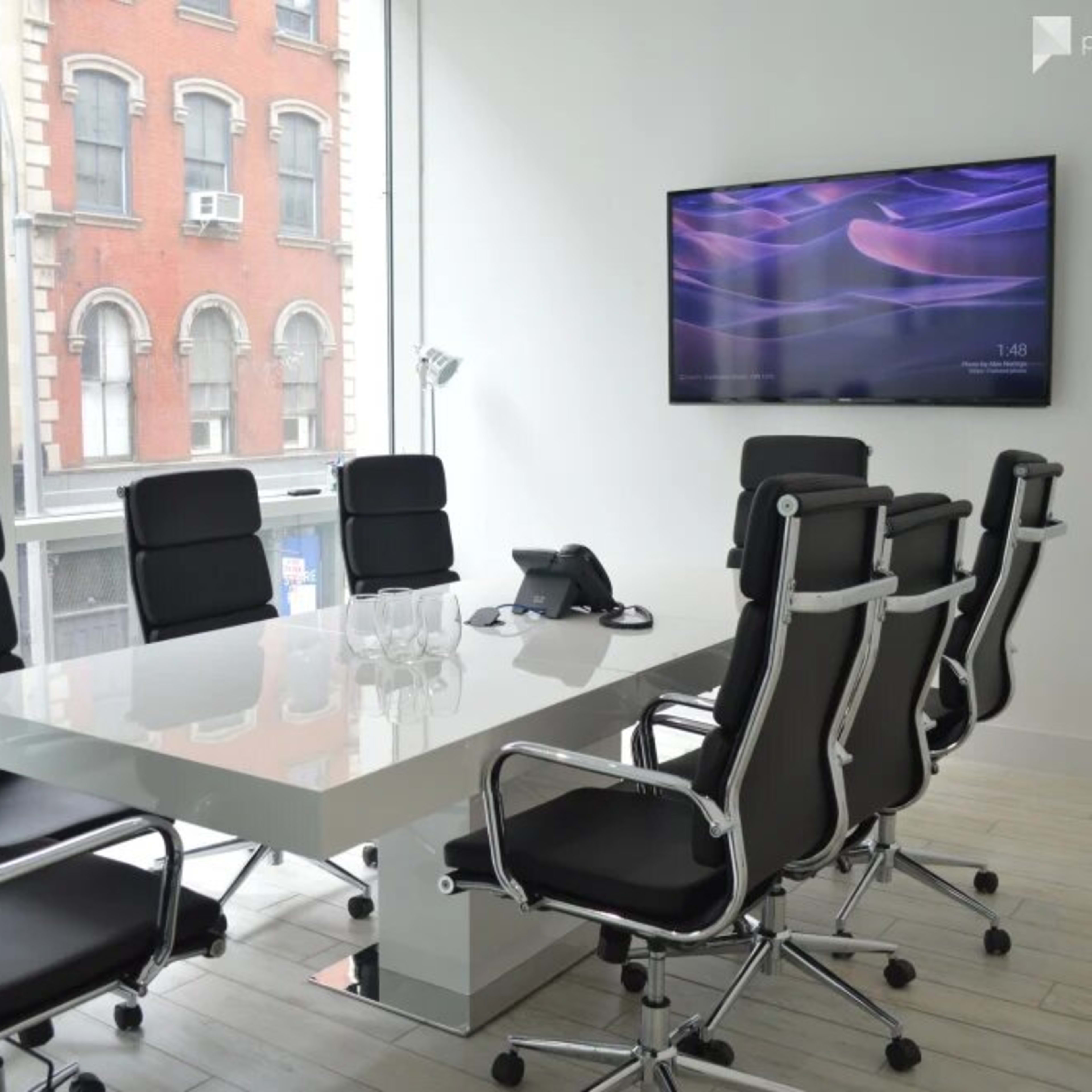 A modern conference room features a large white table surrounded by black ergonomic chairs, with a wall-mounted screen displaying time in the background.