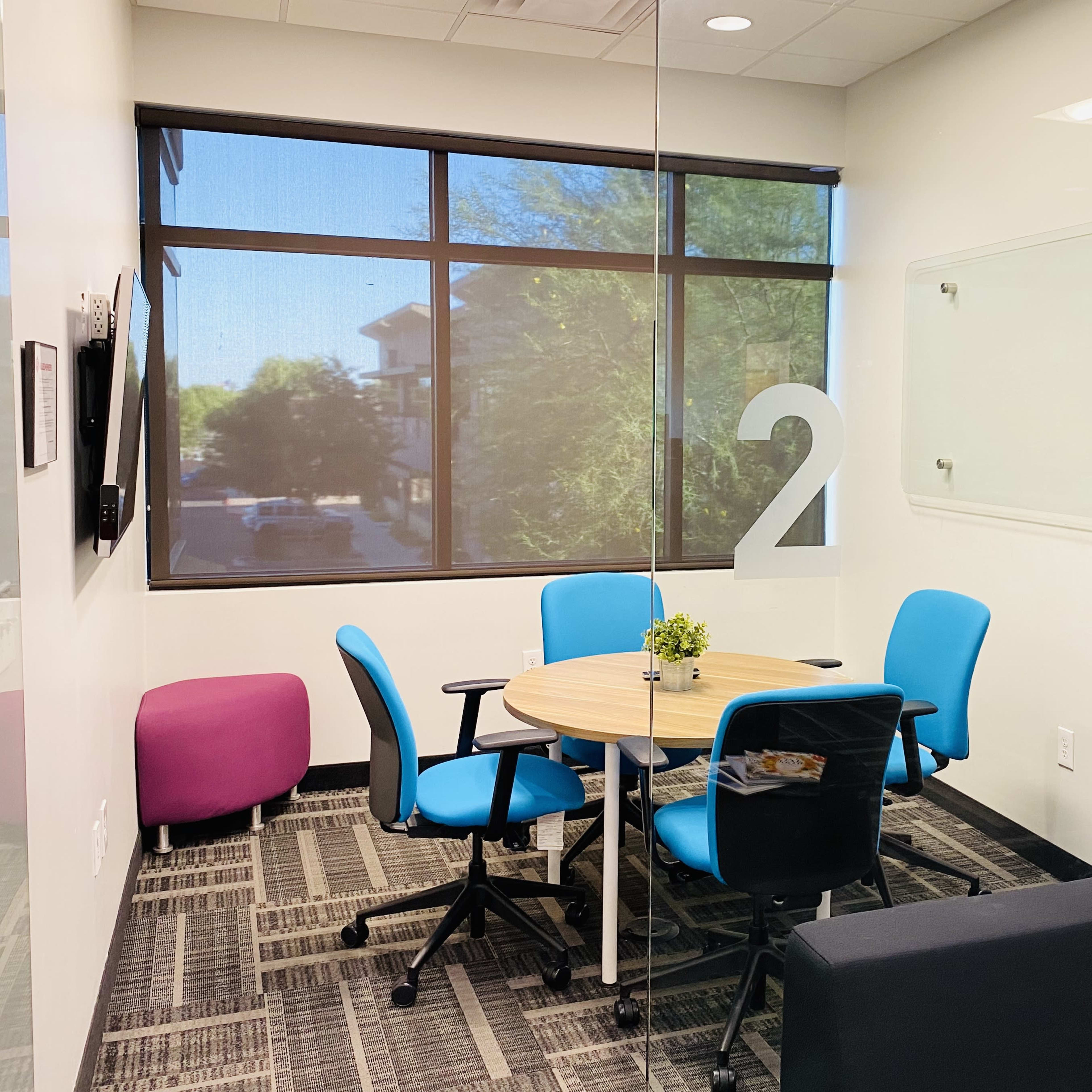 A small meeting room with a round table and four chairs, a potted plant on the table, and a large window letting in natural light.