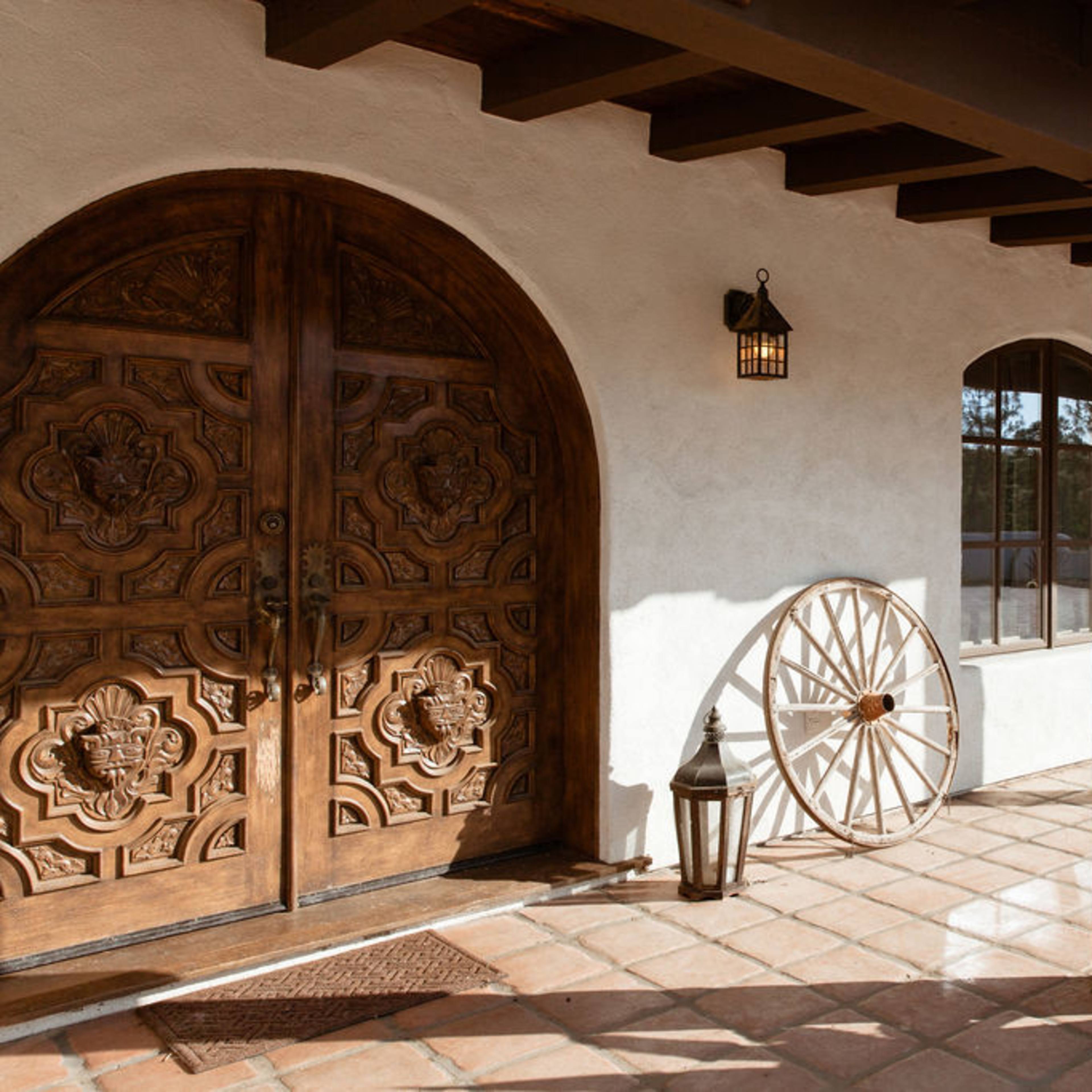 A rustic entrance with a large intricately carved wooden door, a vintage wagon wheel, a potted succulent, and a wooden bench under a tiled roof.