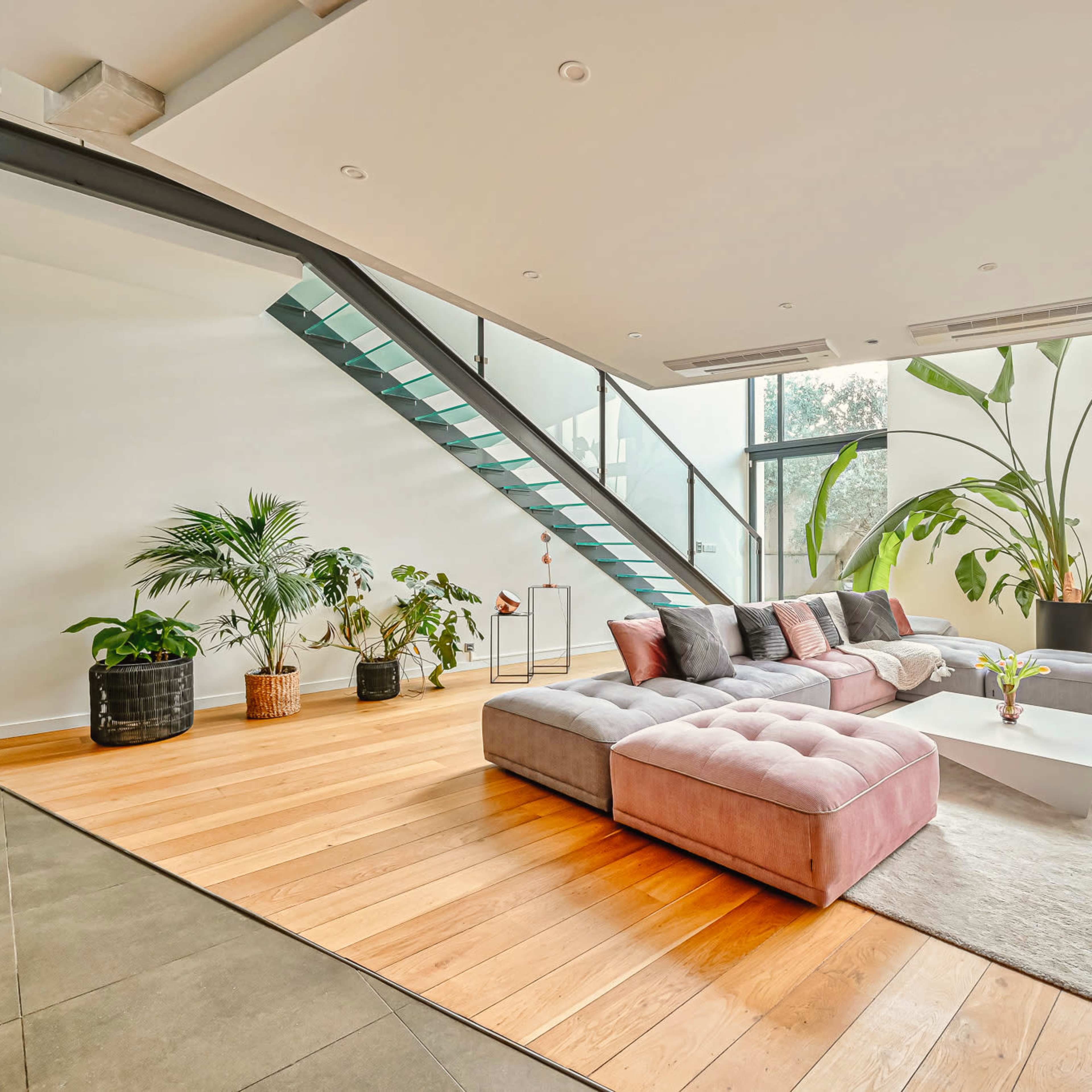 A spacious living room features a modern sectional sofa, large windows, and a staircase with a glass railing.