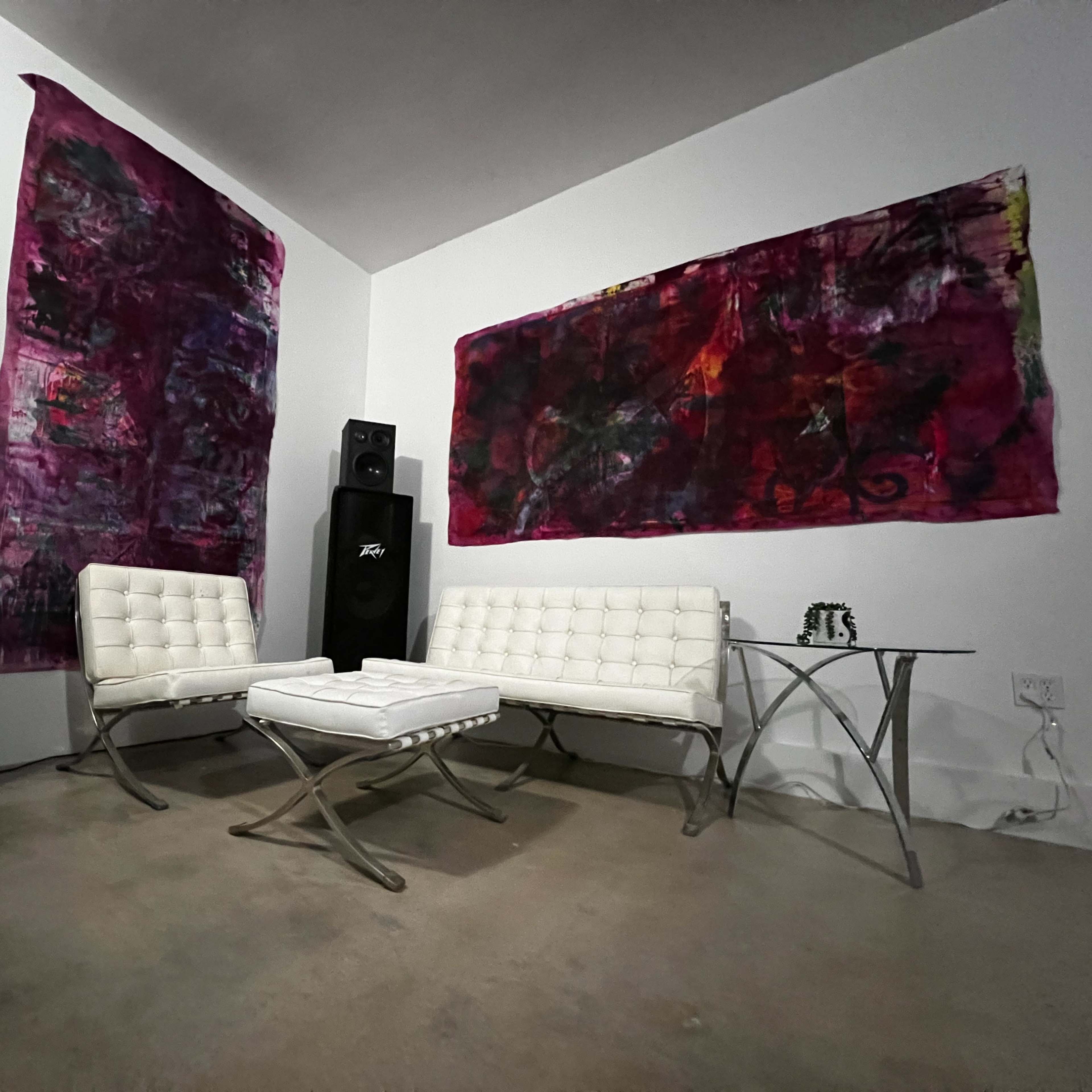 A minimalist seating area with two white chairs and a coffee table against a background of large, colorful abstract paintings and a black speaker.